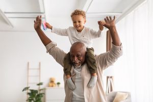 Adult carrying smiling child on shoulders in a bright living room, both with arms raised Adult carrying smiling child on shoulders in a bright living room, both with arms raised