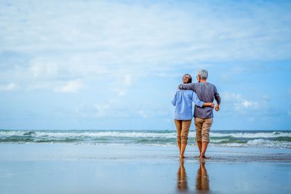 Two people standing arm-in-arm on a beach, facing the ocean under a bright blue sky. Two people standing arm-in-arm on a beach, facing the ocean under a bright blue sky.