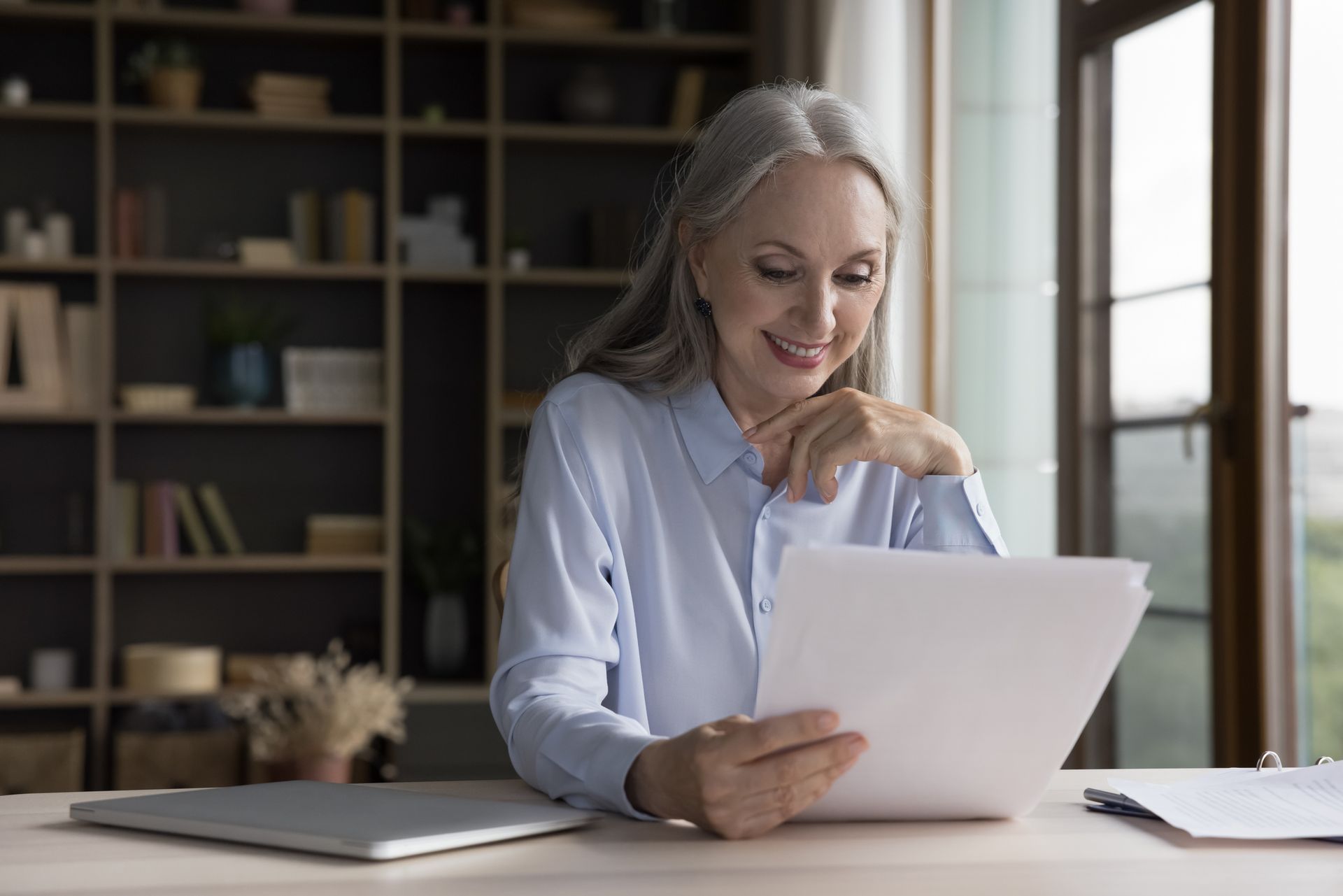 Smiling older woman with gray hair reads papers at a desk in a home office.