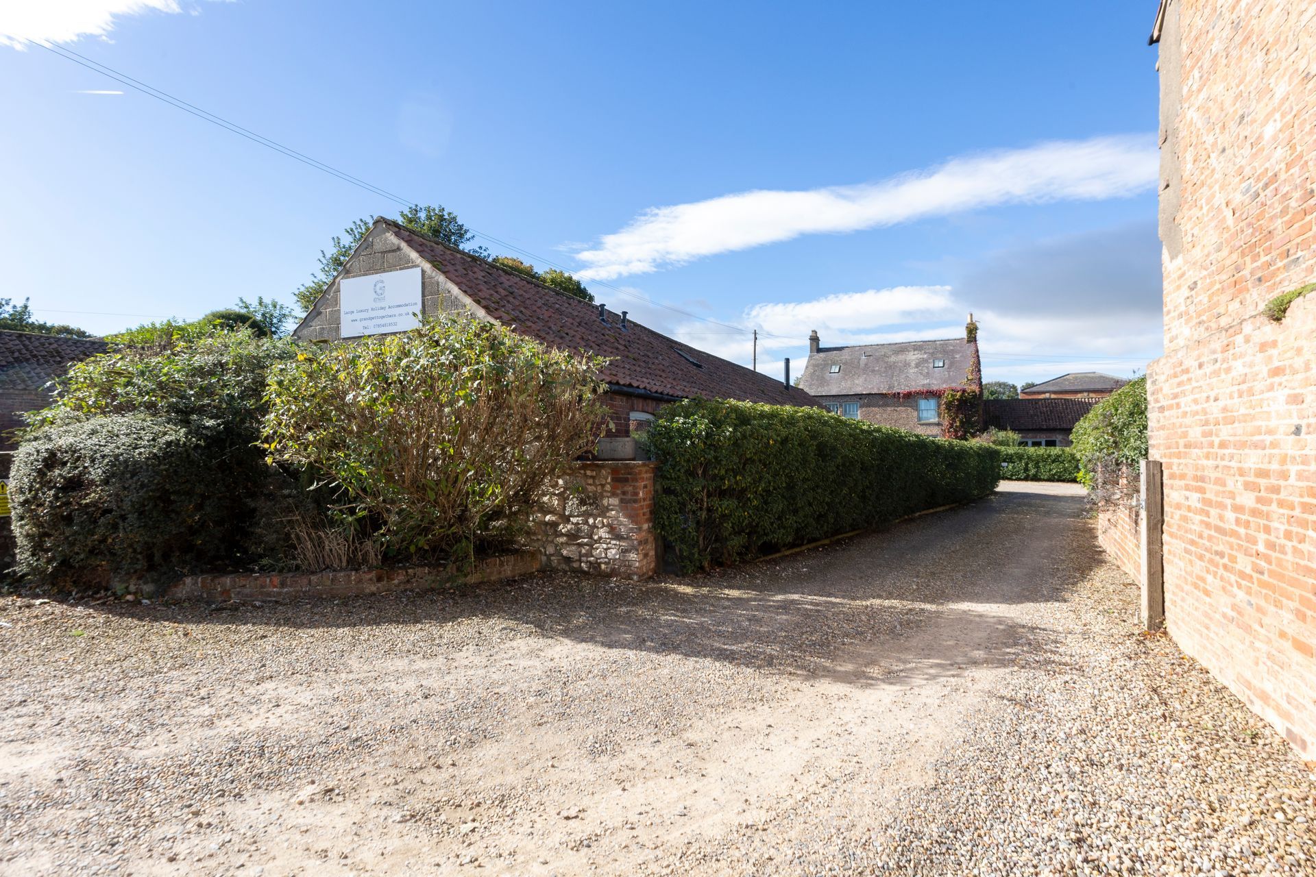 Gravel driveway leads toward buildings, flanked by hedges and brick. Blue sky overhead.