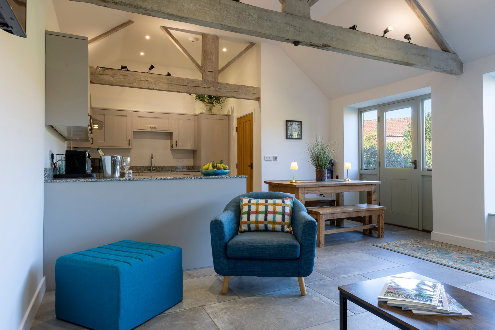 Cozy living space with kitchen, dining area, and teal accent chair, featuring exposed wooden beams and a stone floor.