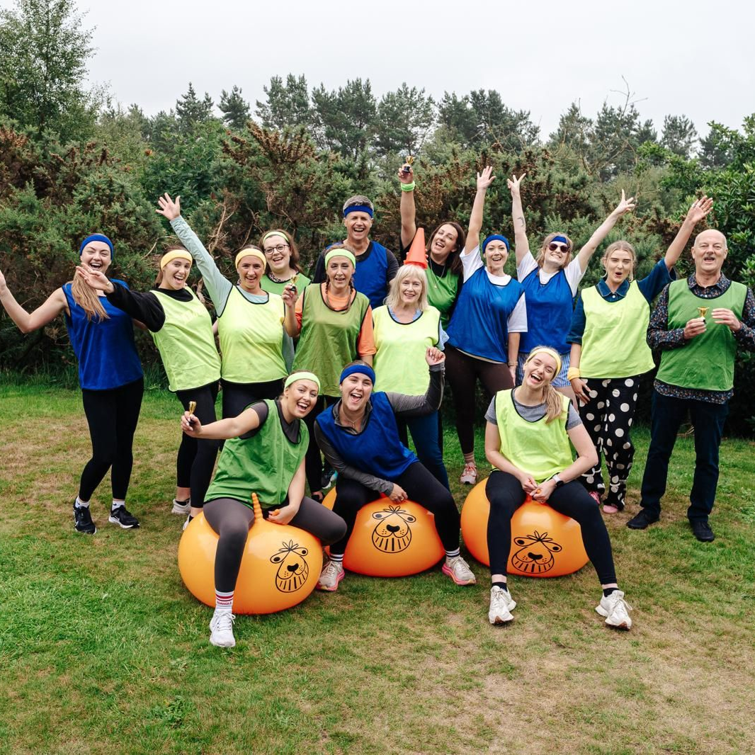 Group of people in vests, bouncing on orange balls, arms raised outdoors.