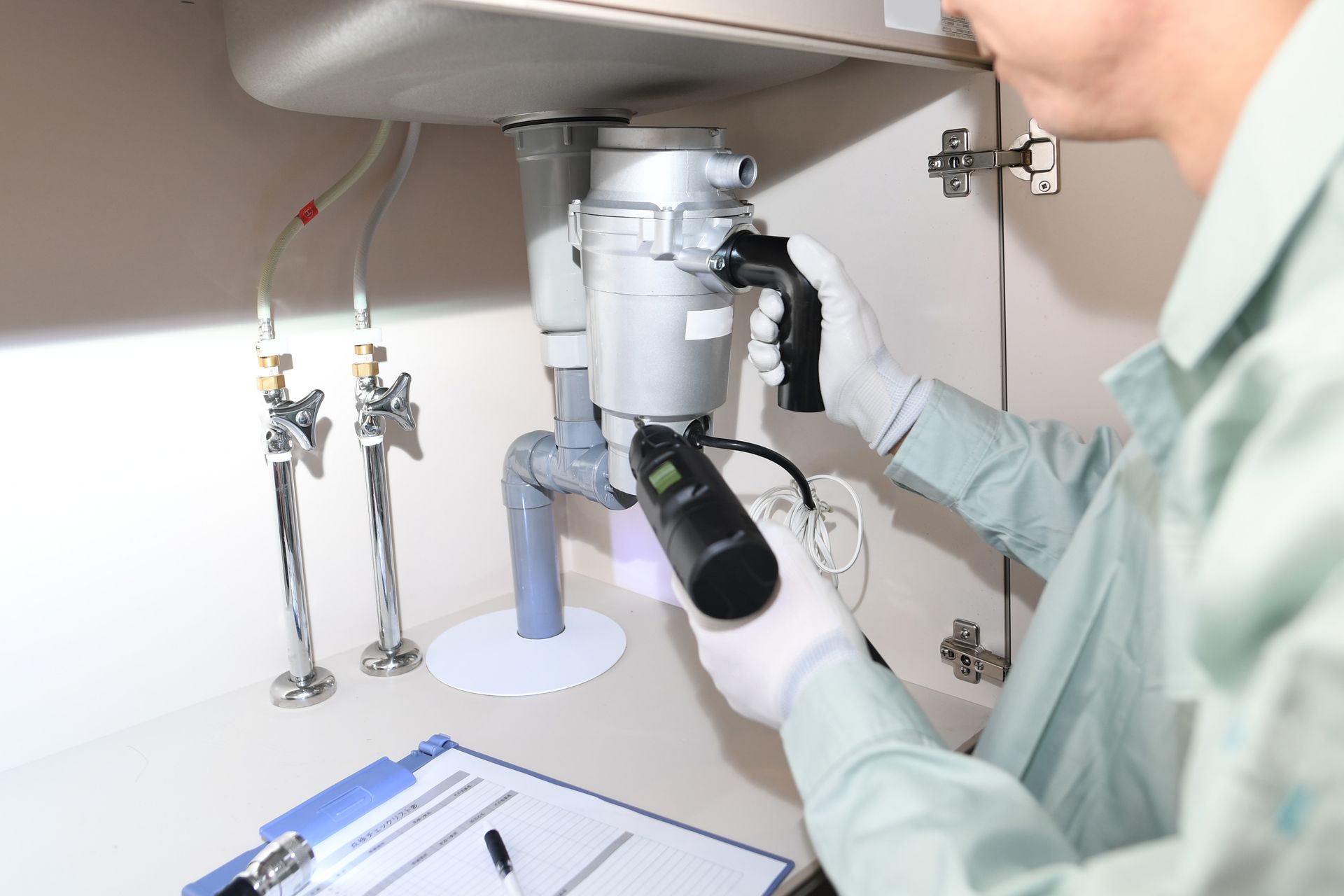 Technician working on a lab instrument under a countertop, with hoses and a clipboard nearby.