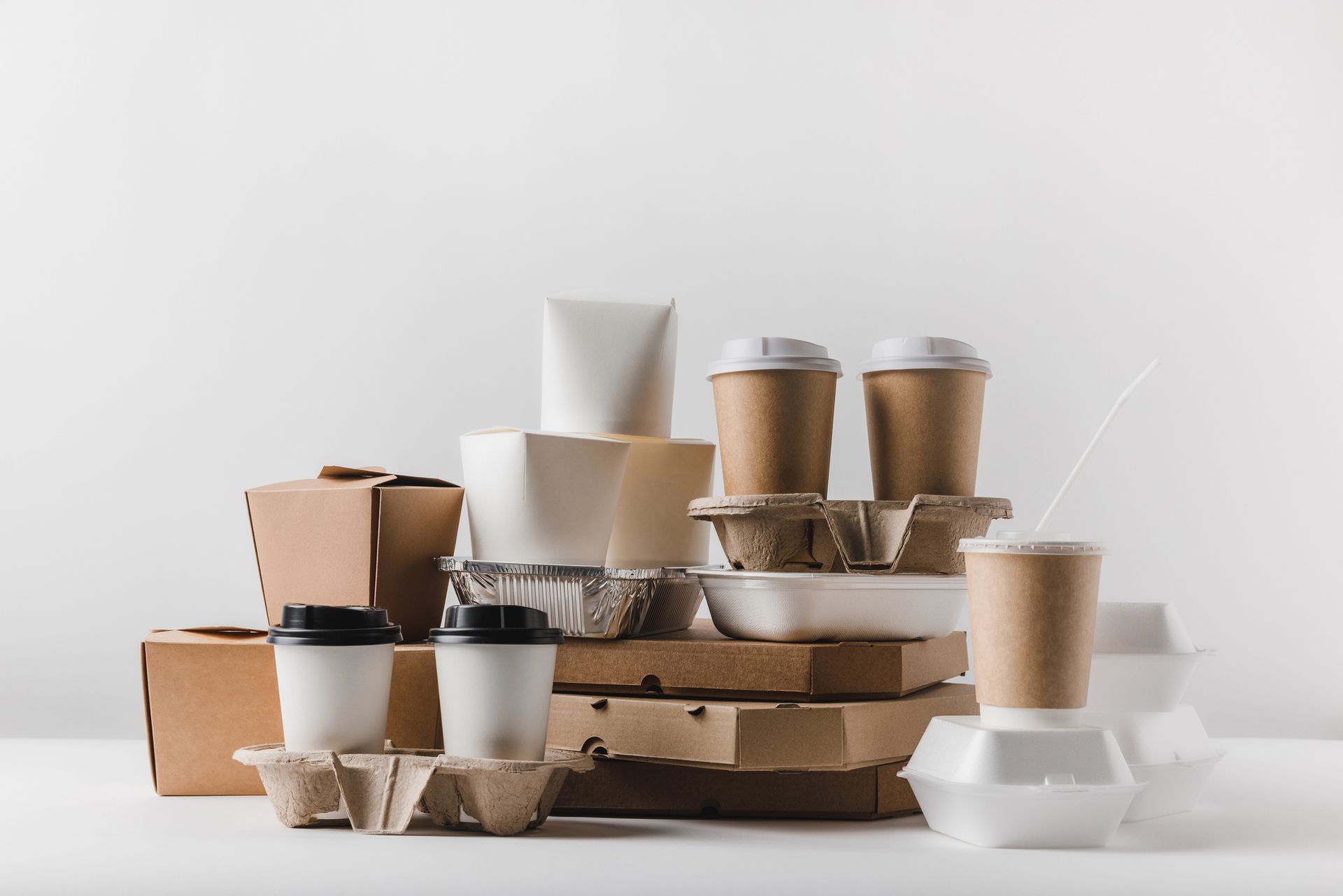 Pila de diversos envases de comida para llevar: cajas de cartón, vasos de café, bandejas de espuma, sobre una superficie blanca.