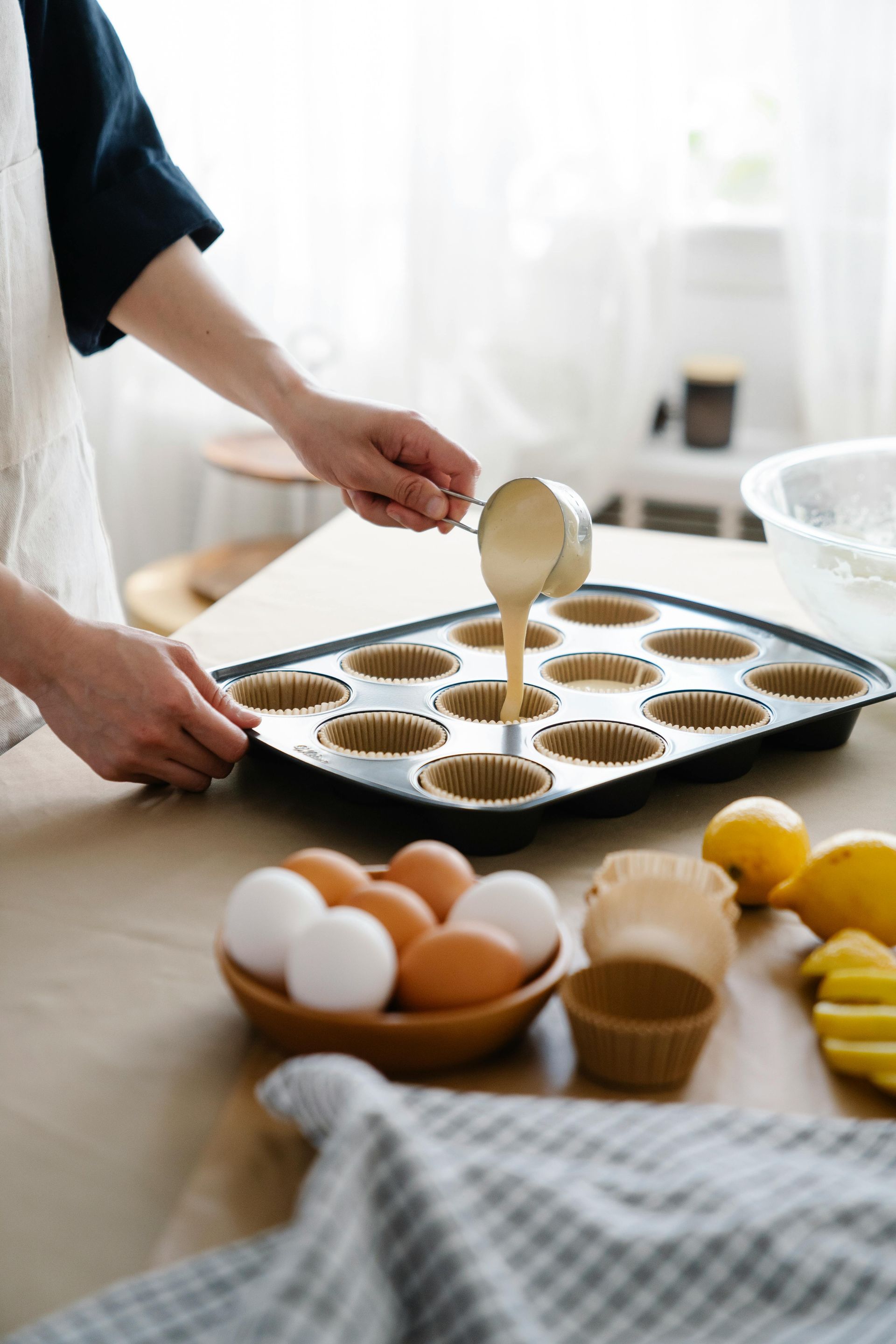 Persona vertiendo masa en un molde para muffins forrado con capsulas de papel; huevos y limones sobre una mesa.