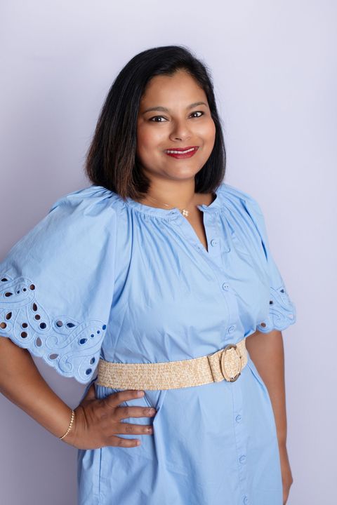 Woman with arms outstretched, smiling in a field with blue sky. She wears a denim jacket over a patterned dress.