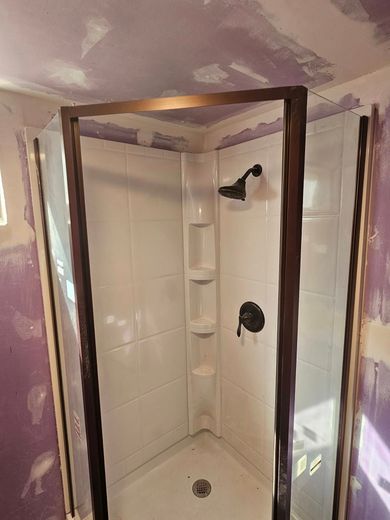 Small tiled shower stall with a dark frame, built-in corner shelves, and unfinished drywall around the opening