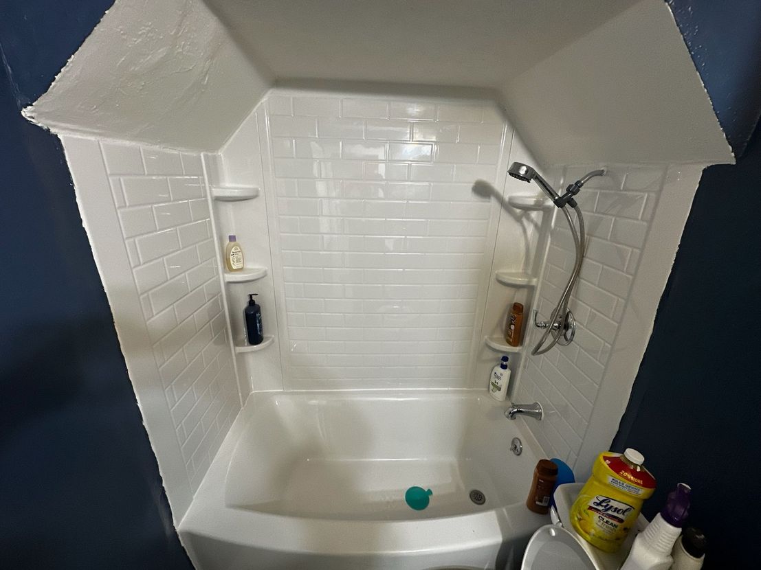 Small white bathtub and shower alcove with tiled walls, toiletries, and cleaning supplies on the ledge