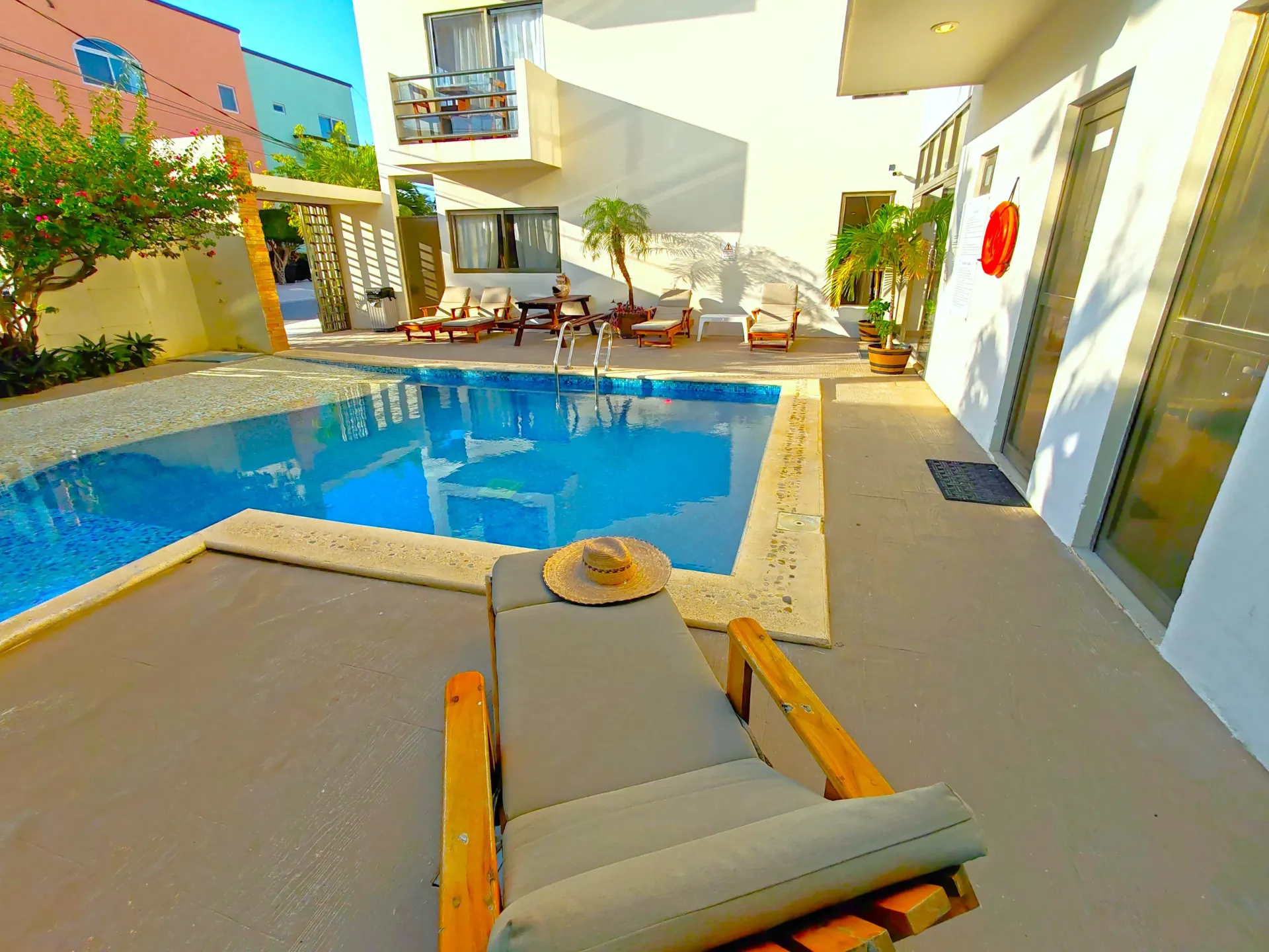 Tumbona con sombrero junto a una piscina en un patio; edificios blancos y cielo azul en el fondo.