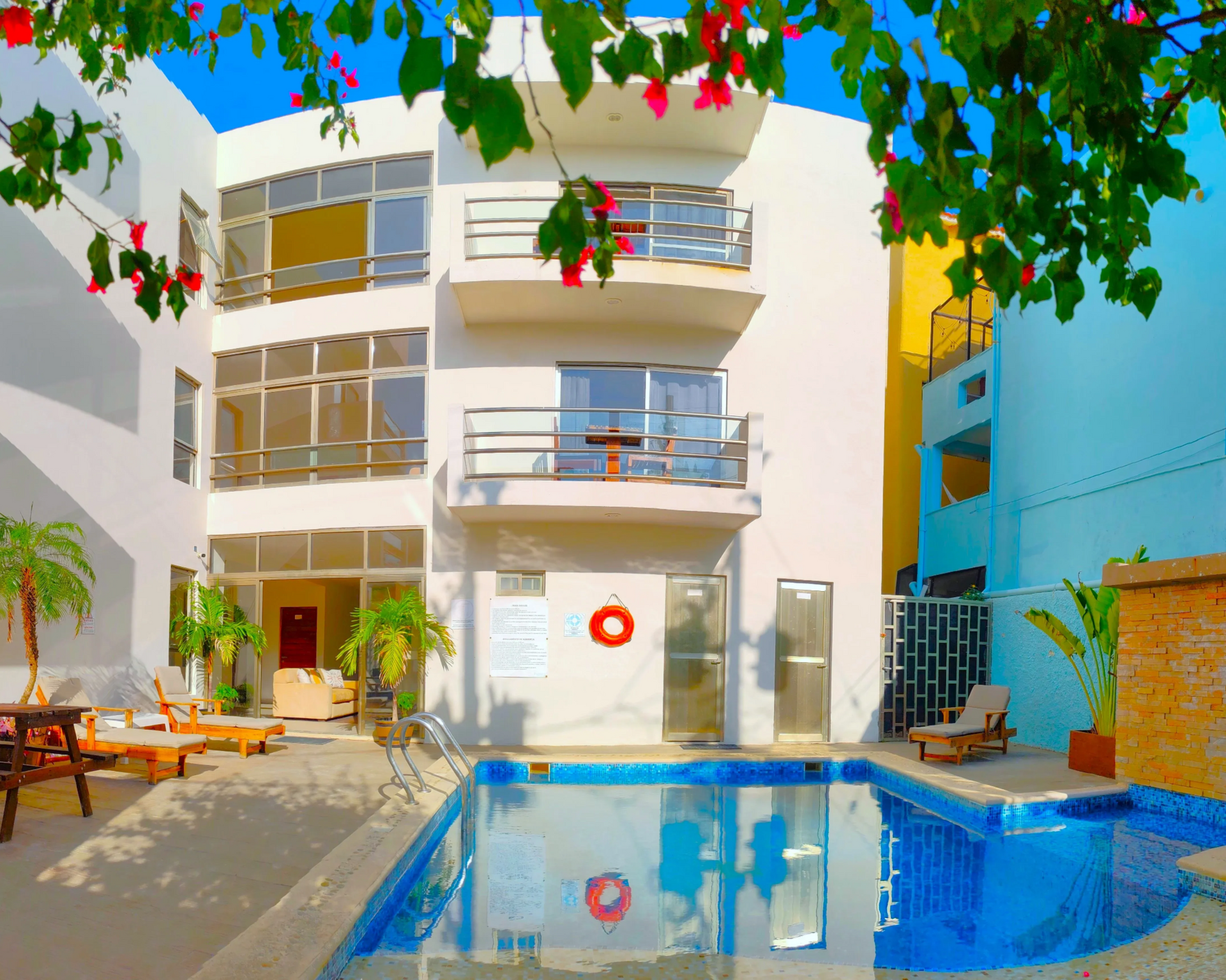 Un pequeño edificio de hotel blanco de tres plantas con balcones que dan a una piscina al aire libre en un día soleado.