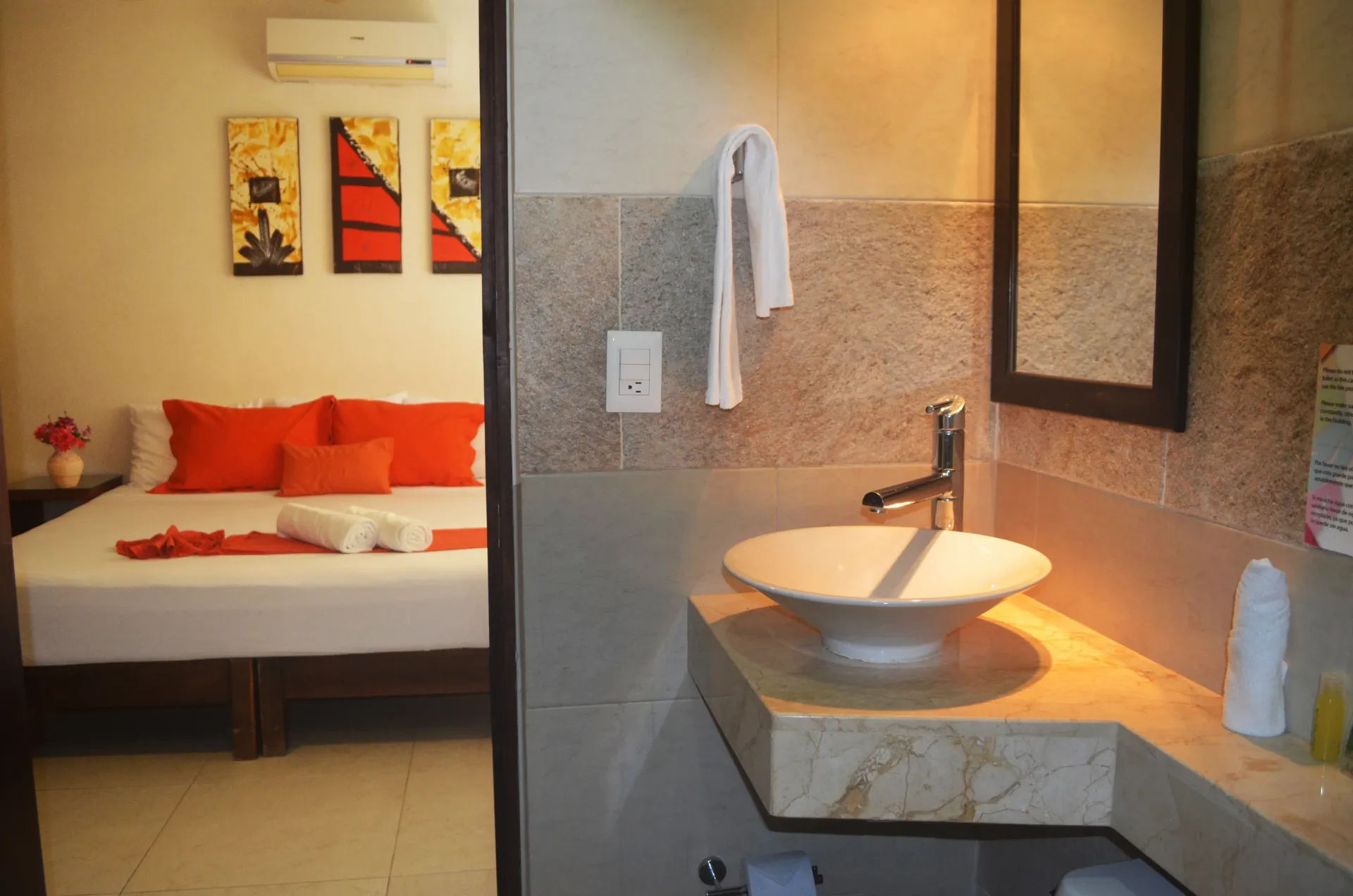 Lavabo del baño y puerta abierta al dormitorio con cama y almohadas naranjas.