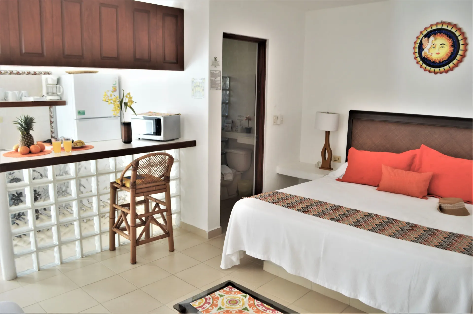 Apartamento tipo estudio con cama, cocina y baño. Incluye un taburete de mimbre, cojines naranjas y un tapiz decorativo.