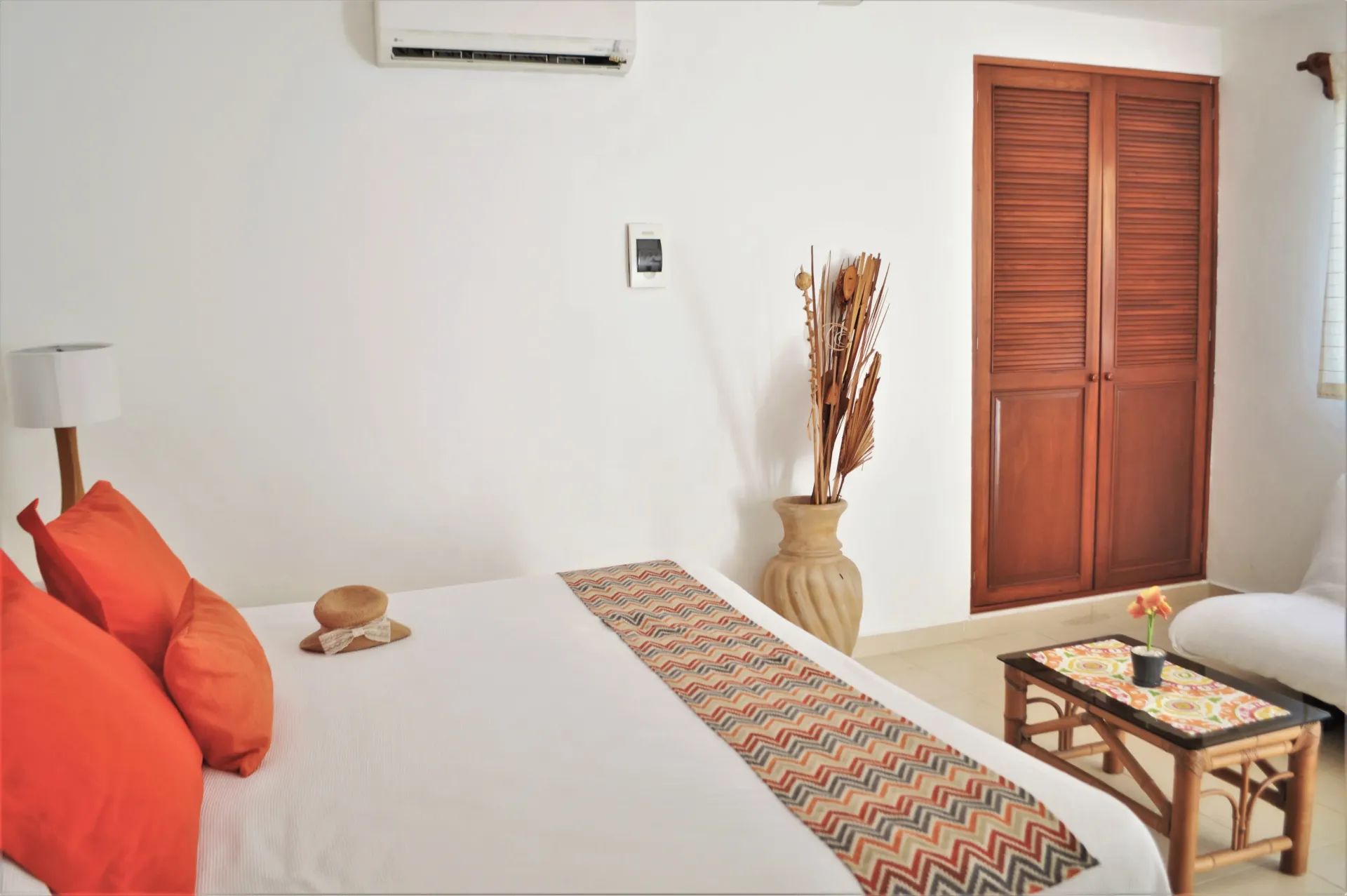 Dormitorio con paredes blancas, cama con almohadas naranjas, puertas de madera y una pequeña mesa.