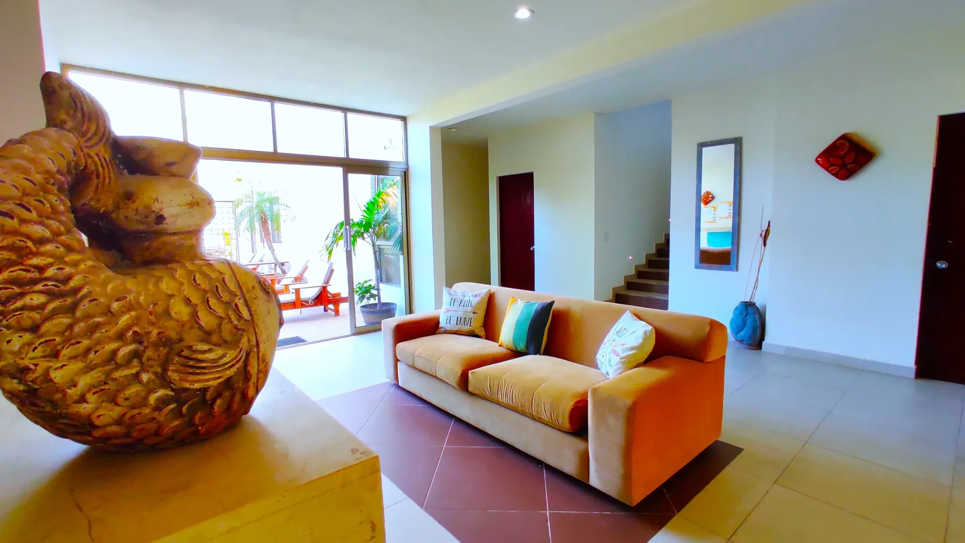 Sala de estar con sofá naranja, puerta abierta a patio, escaleras, objetos decorativos.