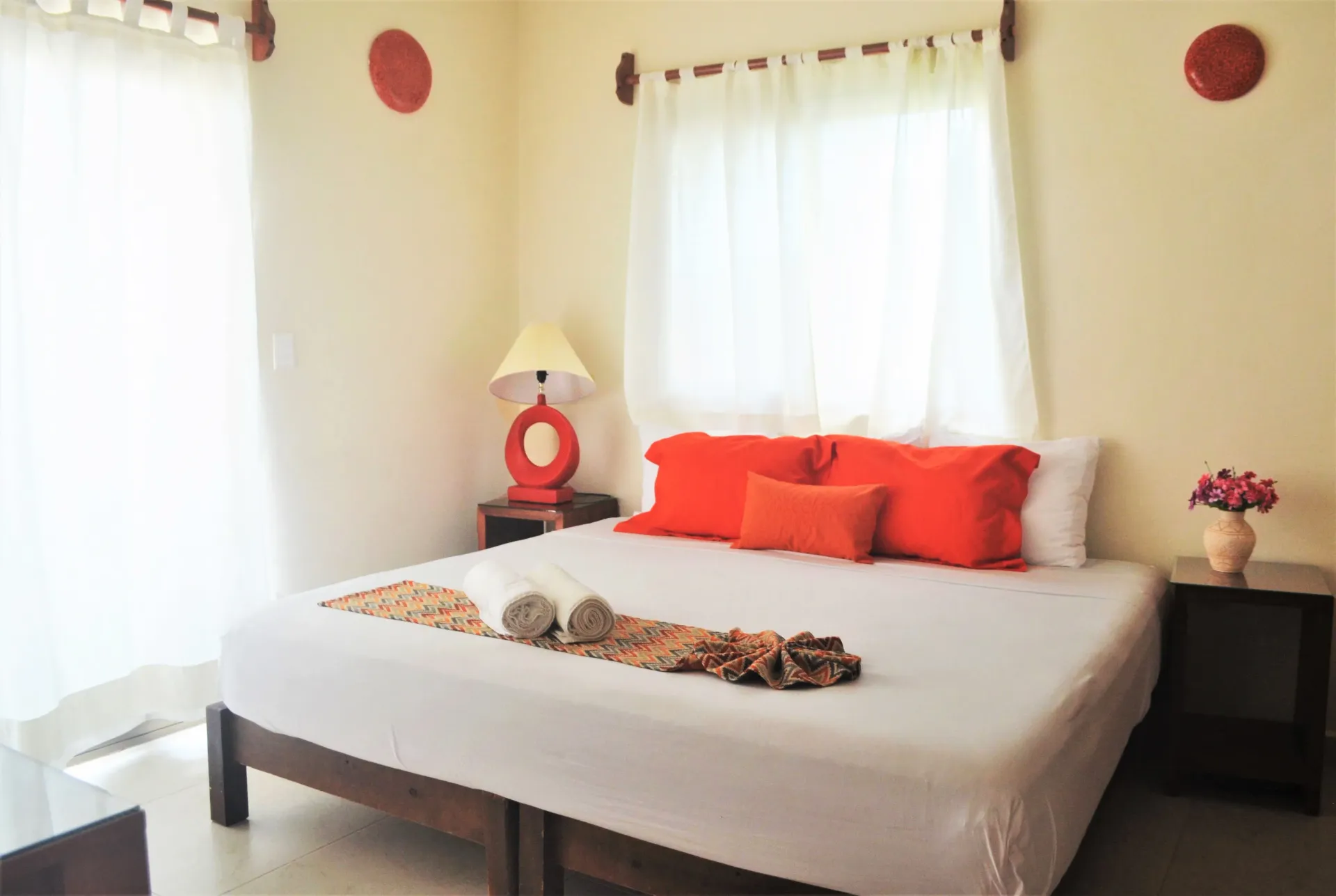 Dormitorio con cama blanca, almohadas naranjas y una ventana con cortinas blancas.