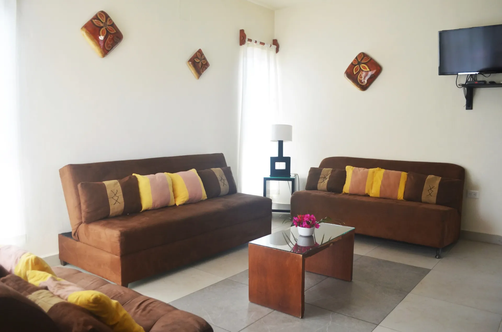 Living room with brown couches, a glass table, and wall art. A mounted TV is visible.