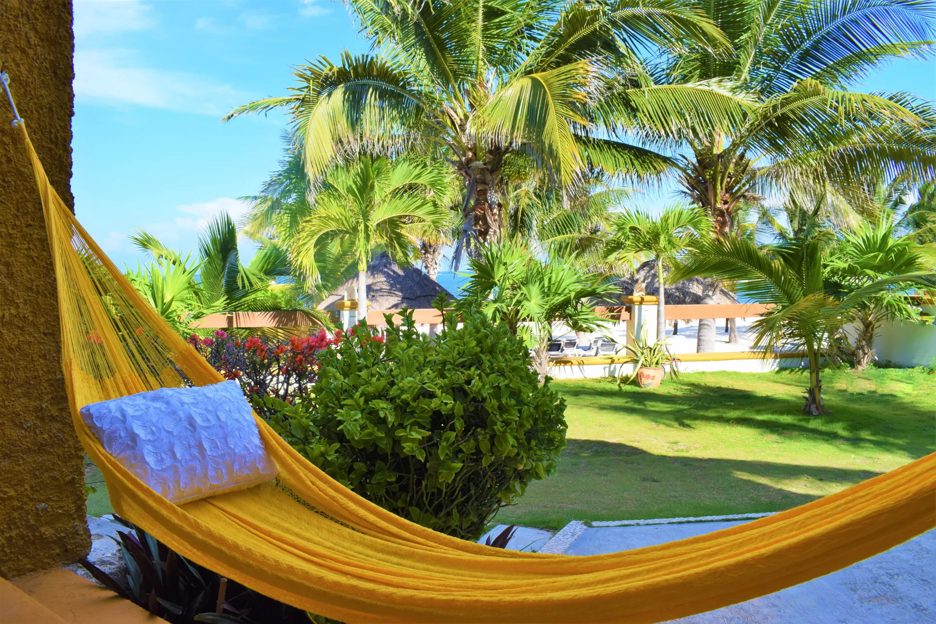 Hamaca amarilla con almohada azul en un porche, con vista a un paisaje tropical con palmeras y césped.