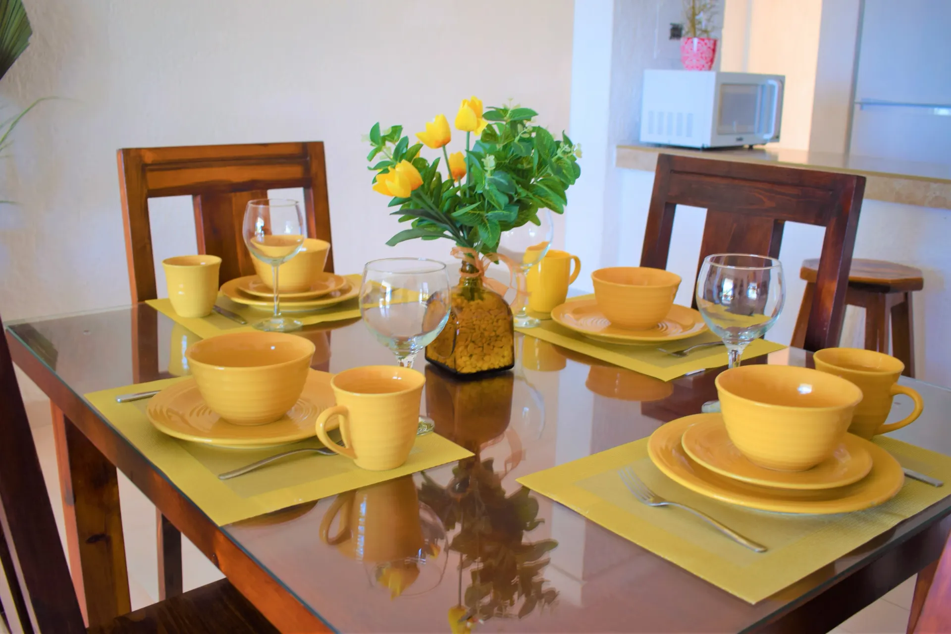 Mesa de comedor con vajilla amarilla, manteles individuales y un centro de mesa floral. Sillas de madera rodean la mesa.