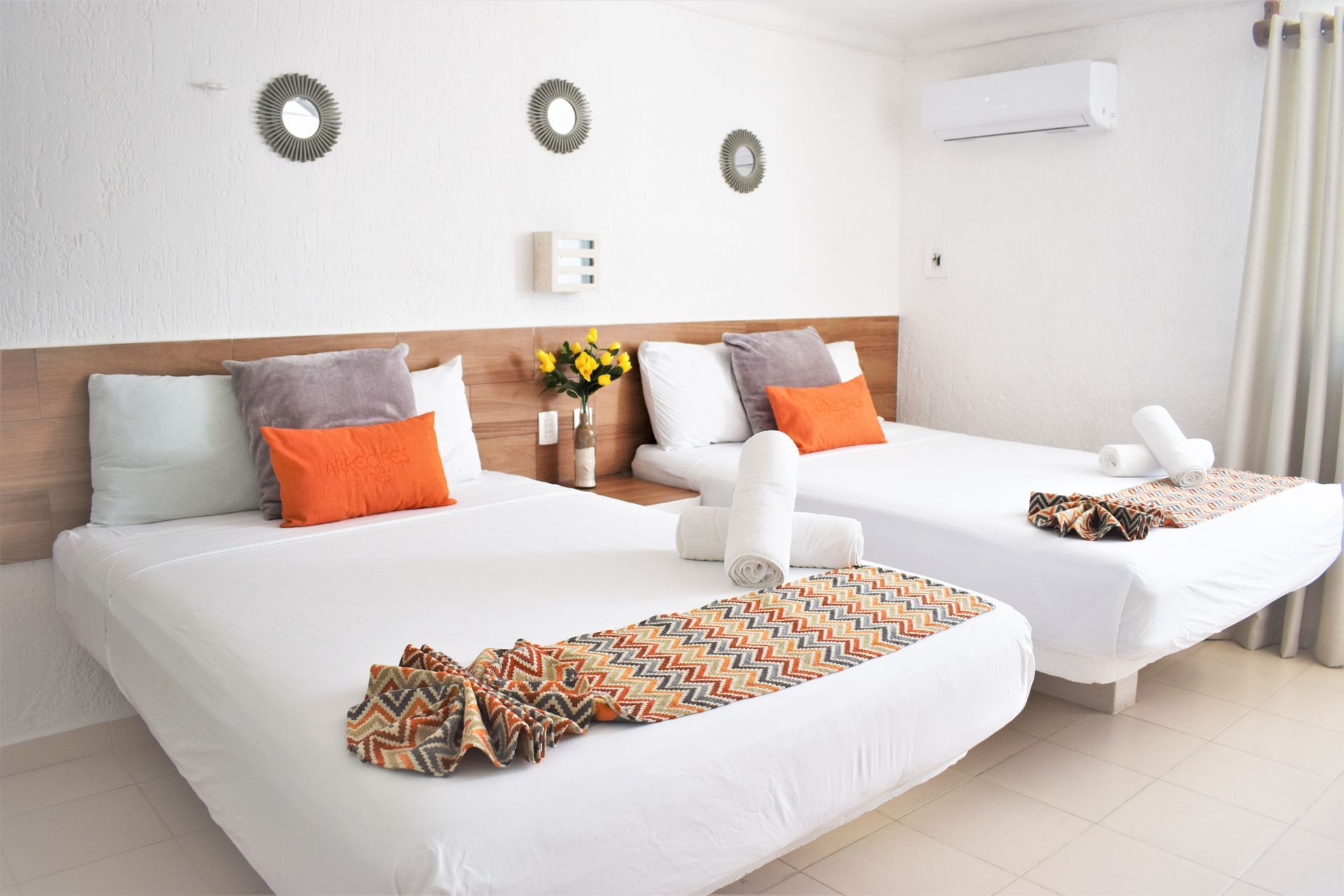 Una habitación de hotel con dos camas, paredes blancas, espejos decorativos y almohadas y mantas de colores.