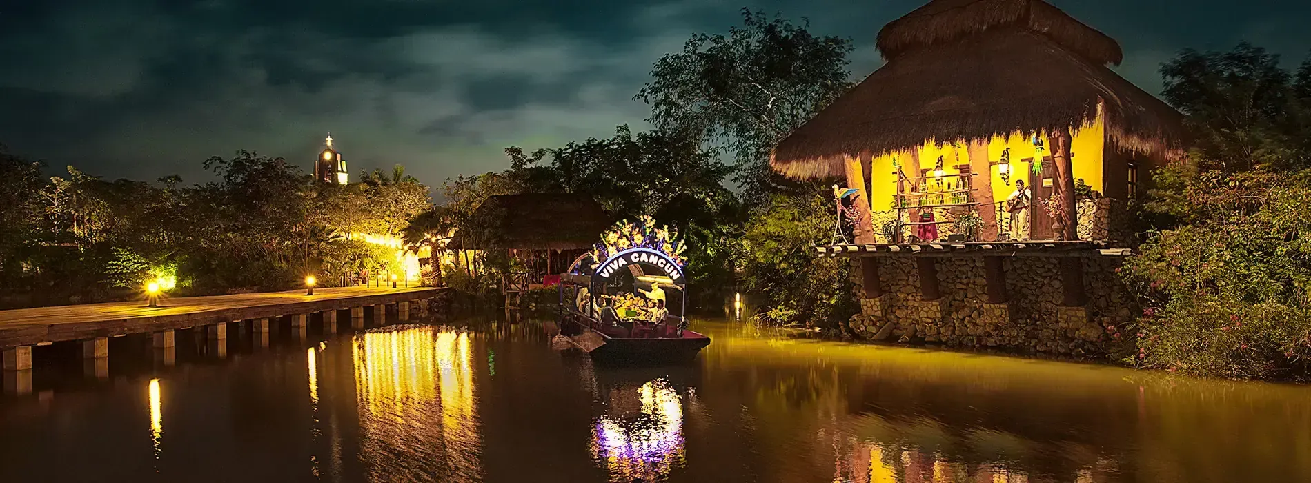 Night scene of illuminated huts and bridge over water; lights reflecting in the water.