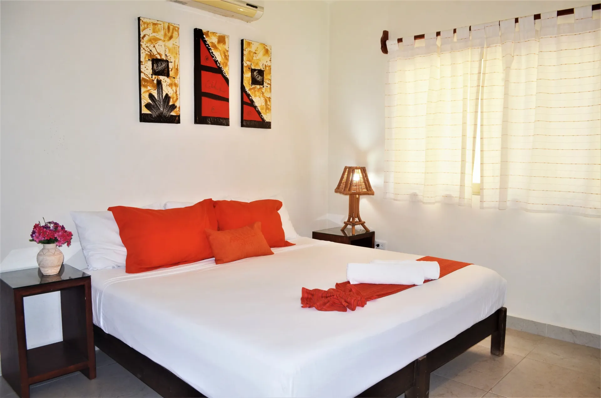 Dormitorio con paredes blancas, cama con almohadas naranjas y obras de arte.
