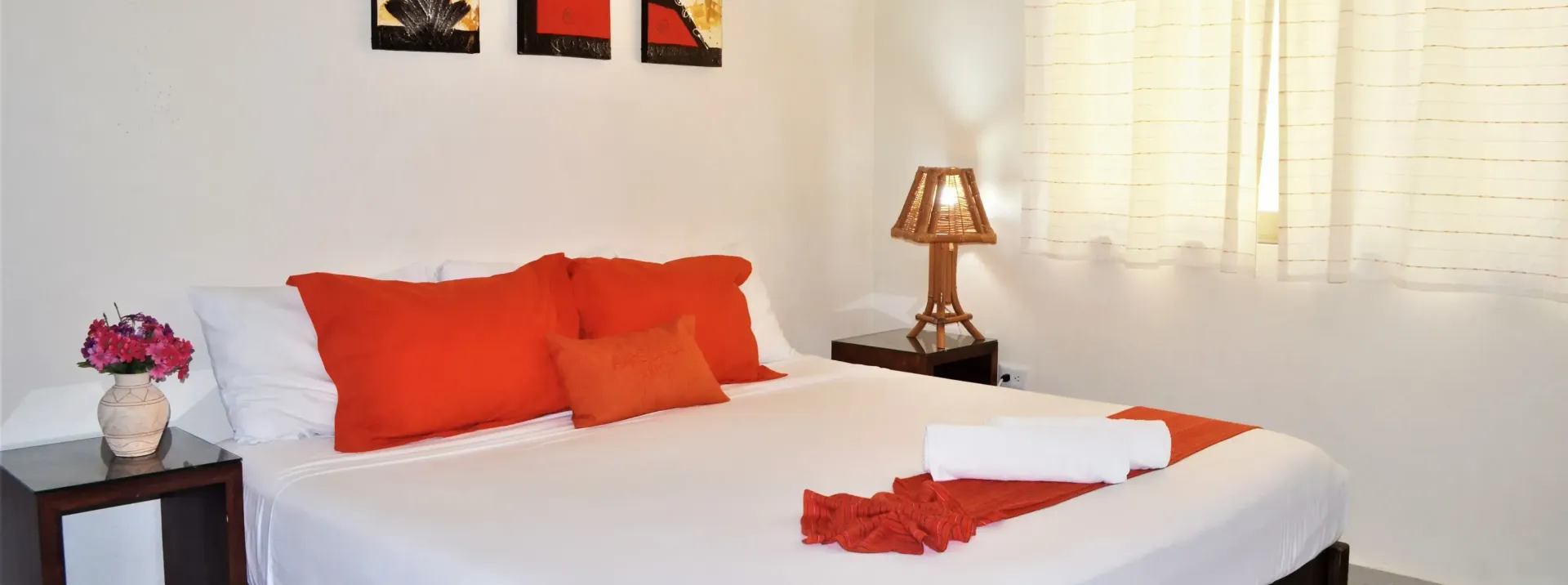 Cama hecha con sábanas blancas,  almohadas naranjas y una lámpara de noche en una habitación luminosa.
