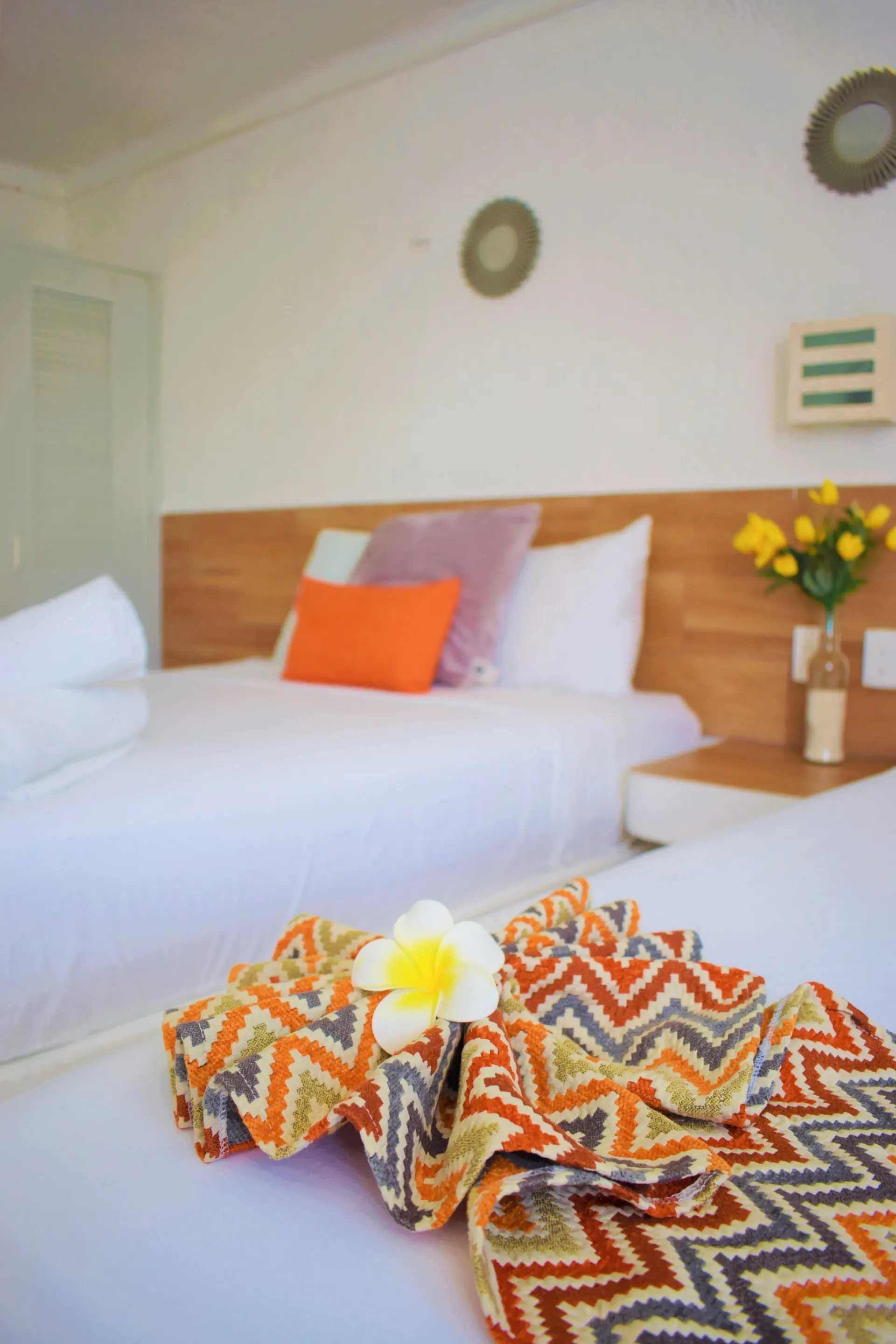 Dos camas con sábanas blancas, almohadas de color naranja y morado y una manta estampada doblada con una flor encima.
