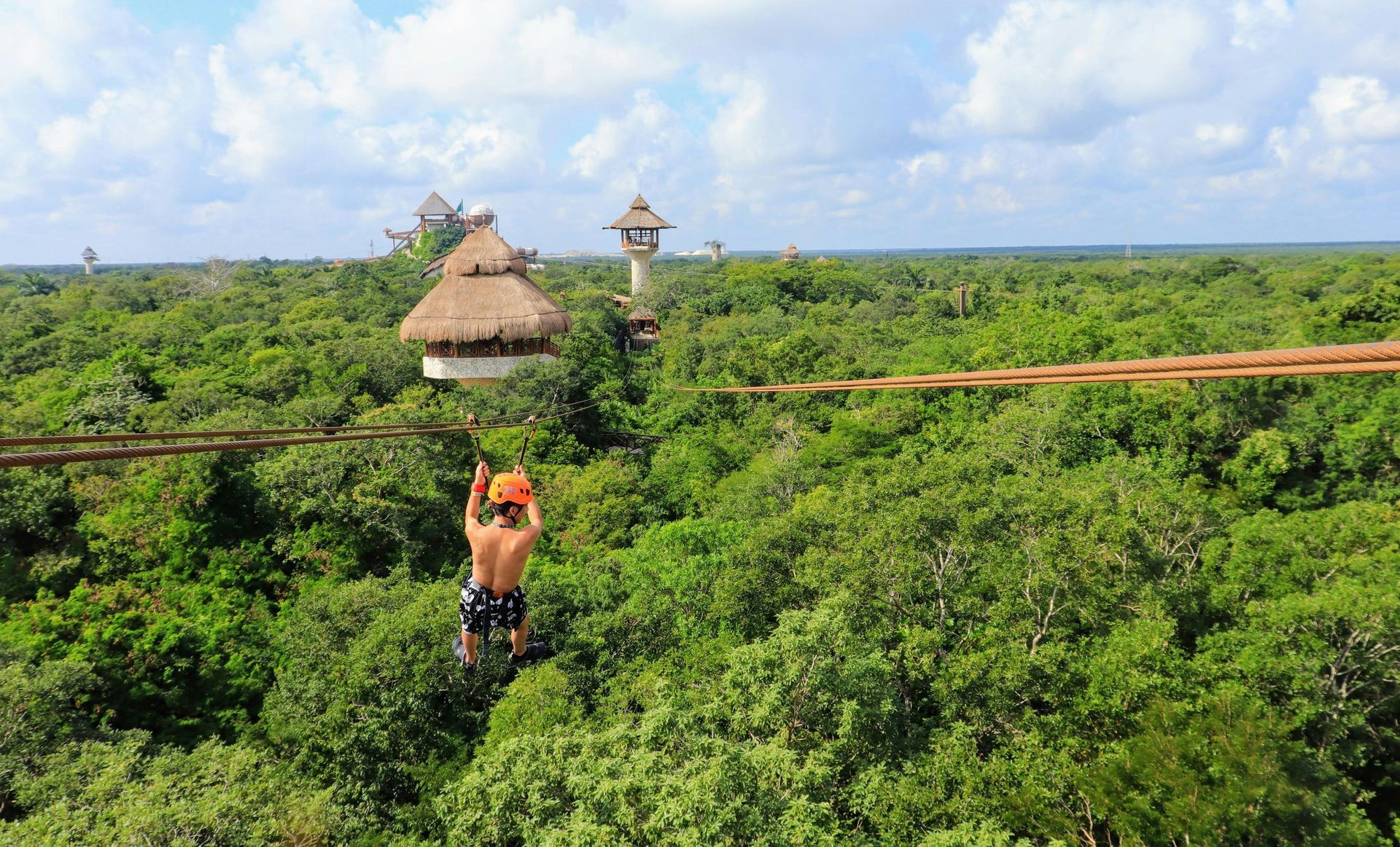 Person ziplining over lush green jungle toward a thatched-roof tower on a sunny day.