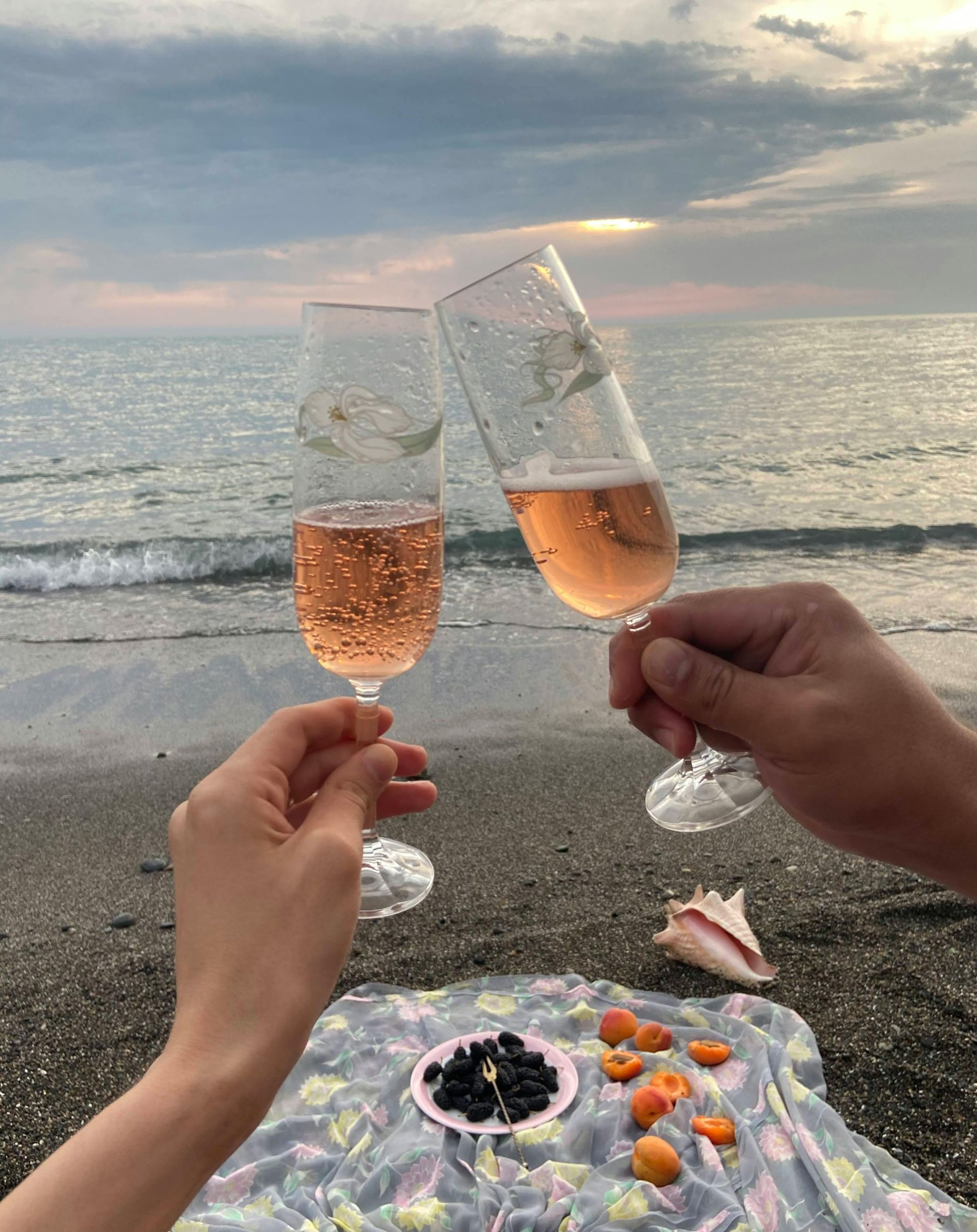 Two hands clinking champagne flutes on a beach at sunset, with a blanket and snacks.