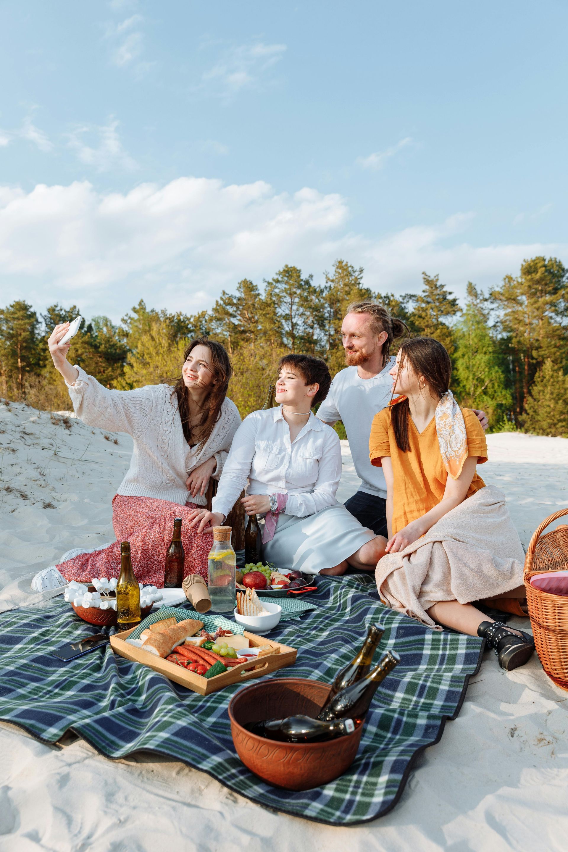 Four friends on a beach picnic; one takes a selfie. Blue blanket, food spread, trees and sky in the background.
