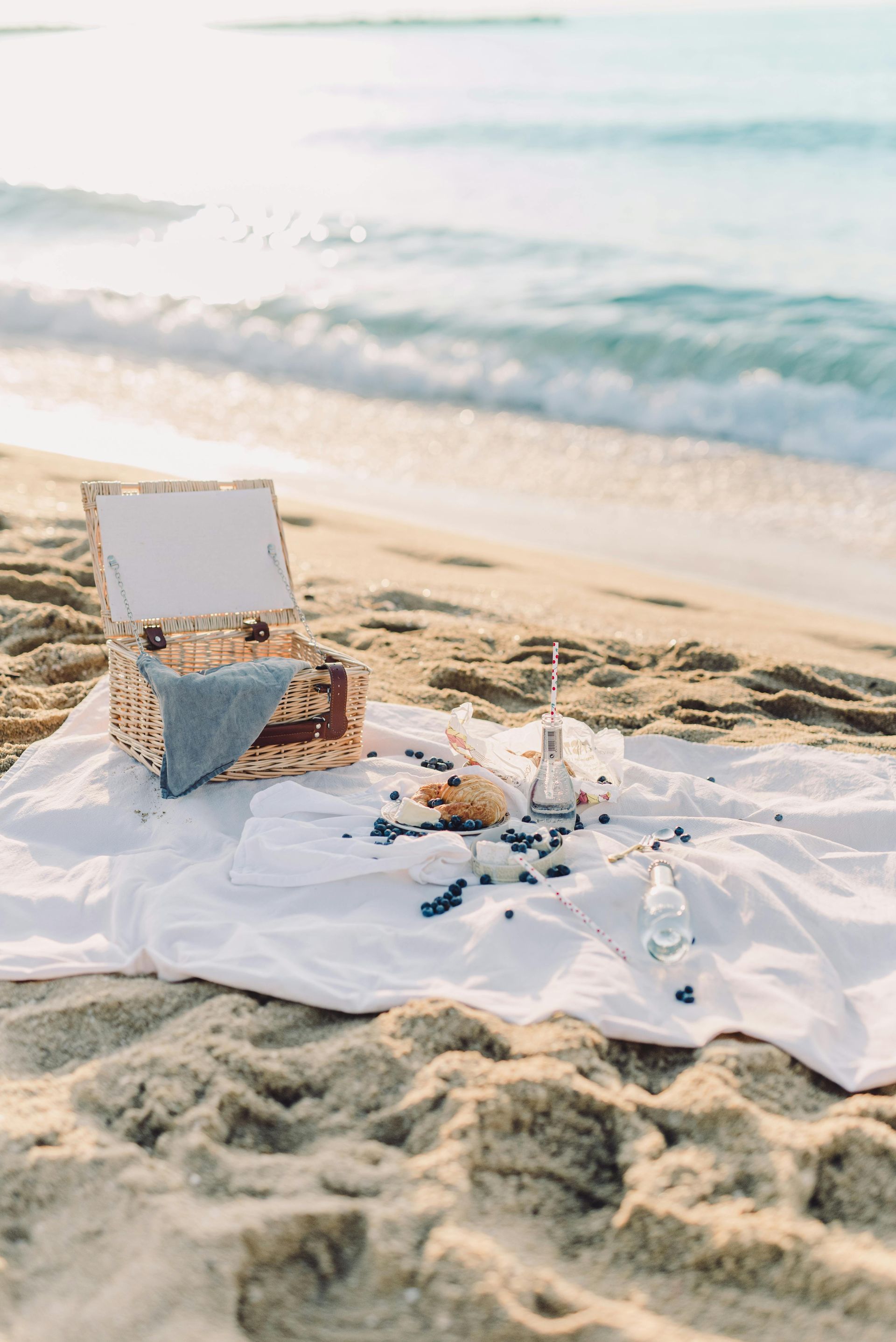 Picnic on the beach: woven basket, white blanket, blueberries, food, and drinks on the sand near the ocean.