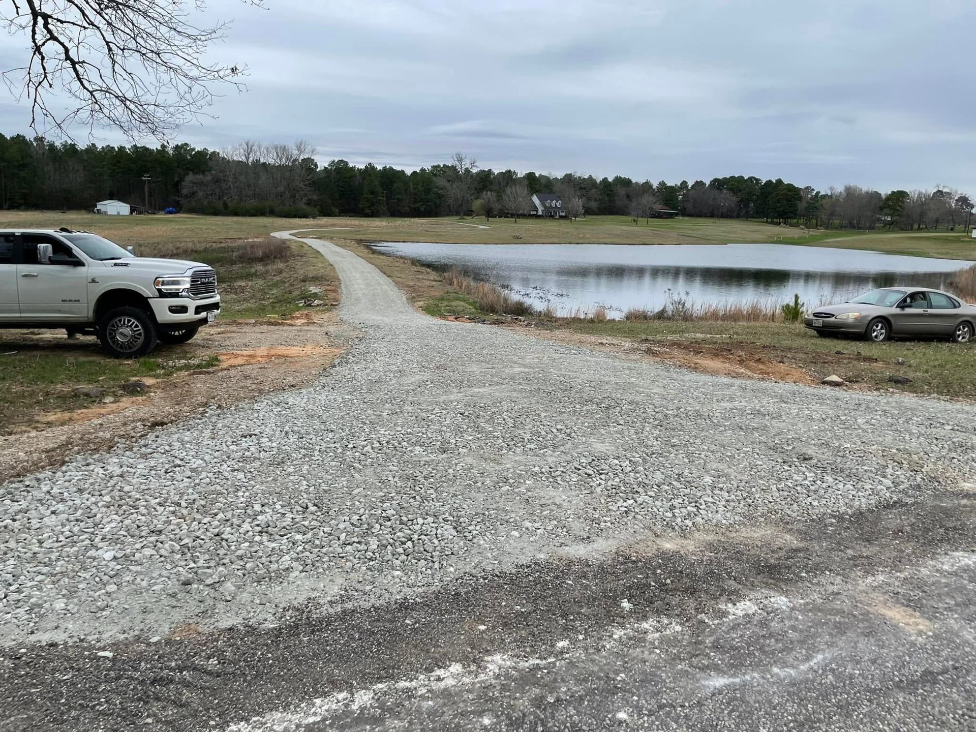 Gravel driveway by a pond with a white truck on the left and a silver car on the right under cloudy skies