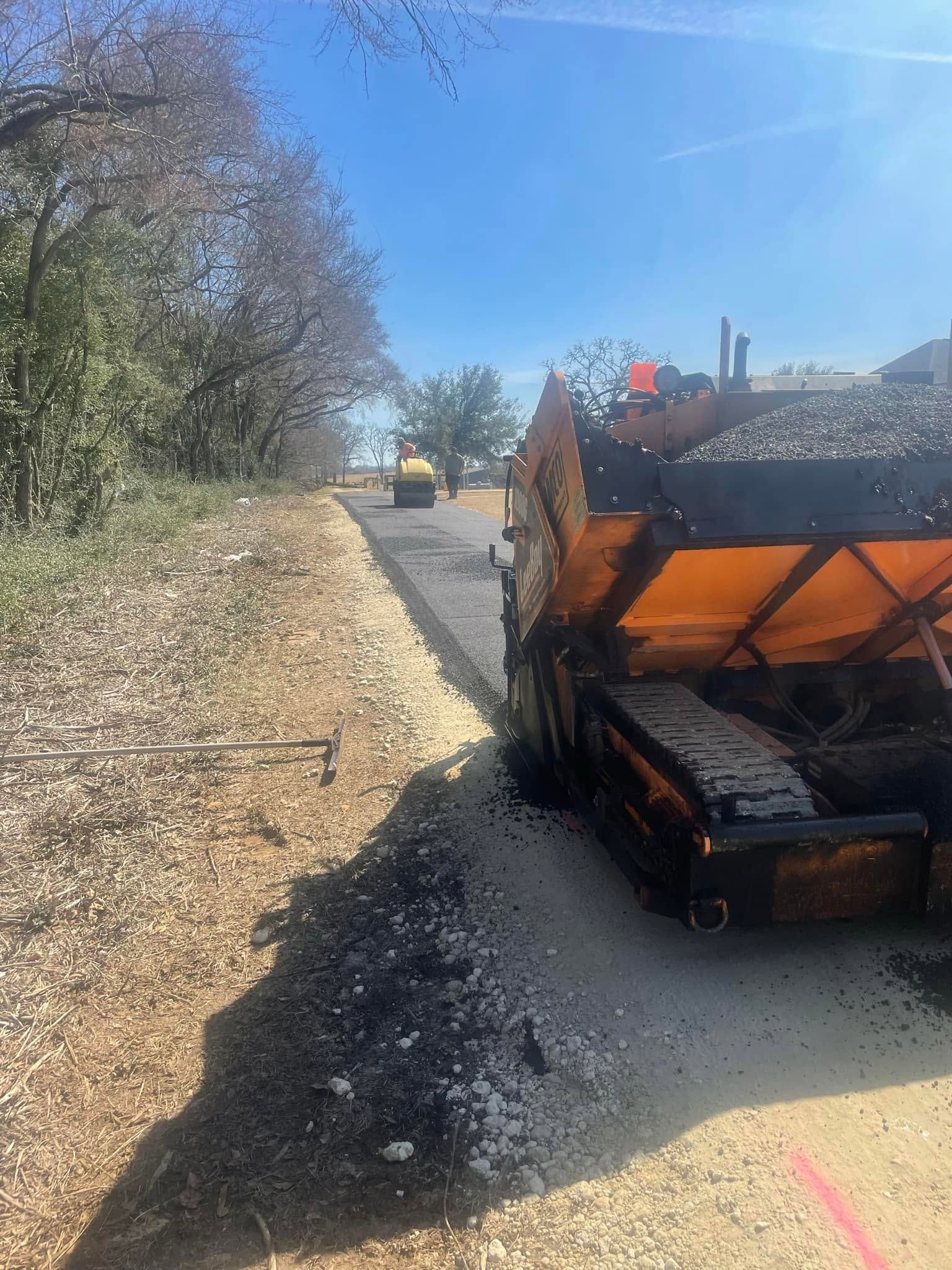 Road paving machine spreading black asphalt on a rural road under a clear blue sky