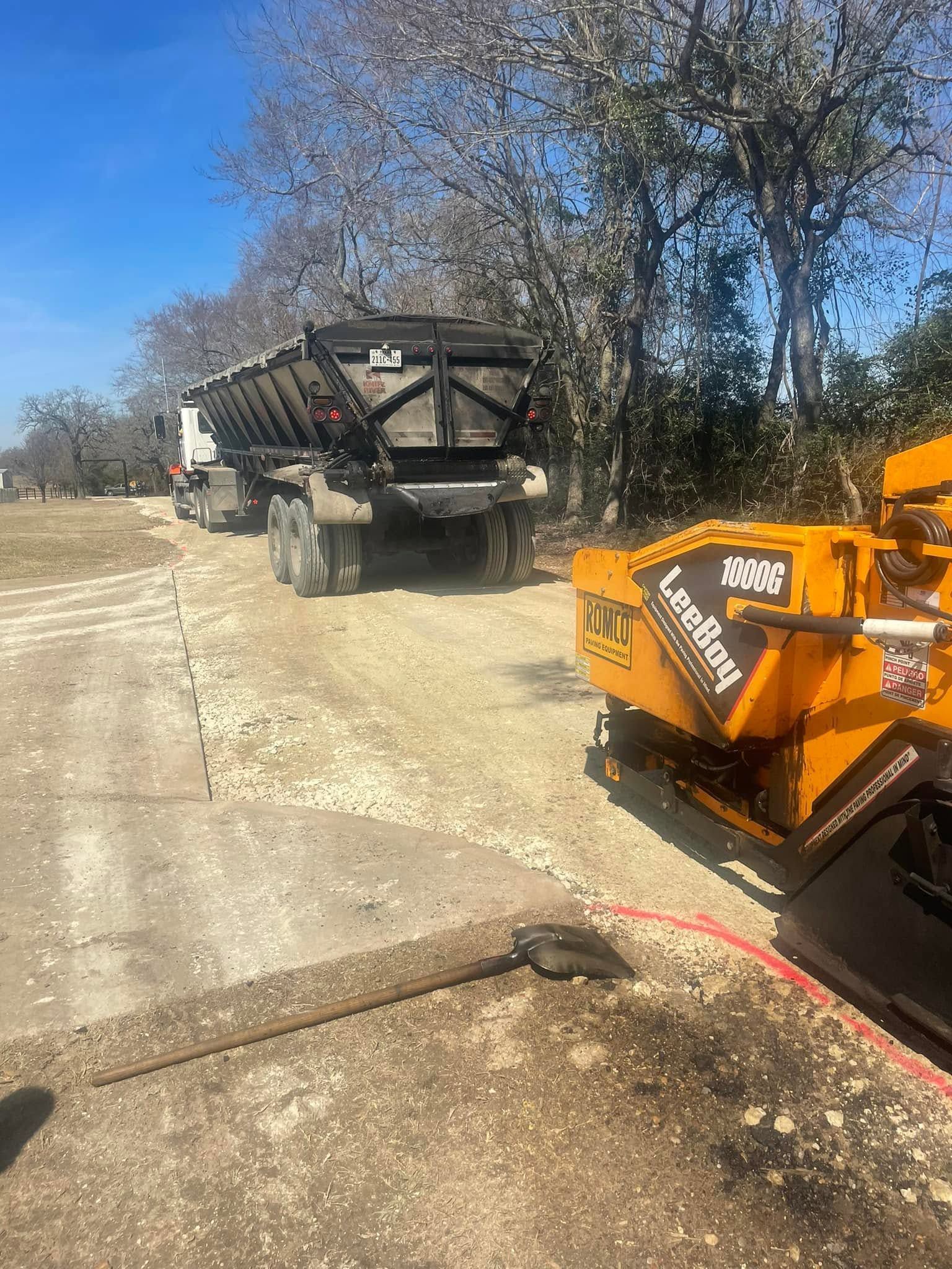 Orange construction equipment beside a dump truck on a gravel roadside under a blue sky
