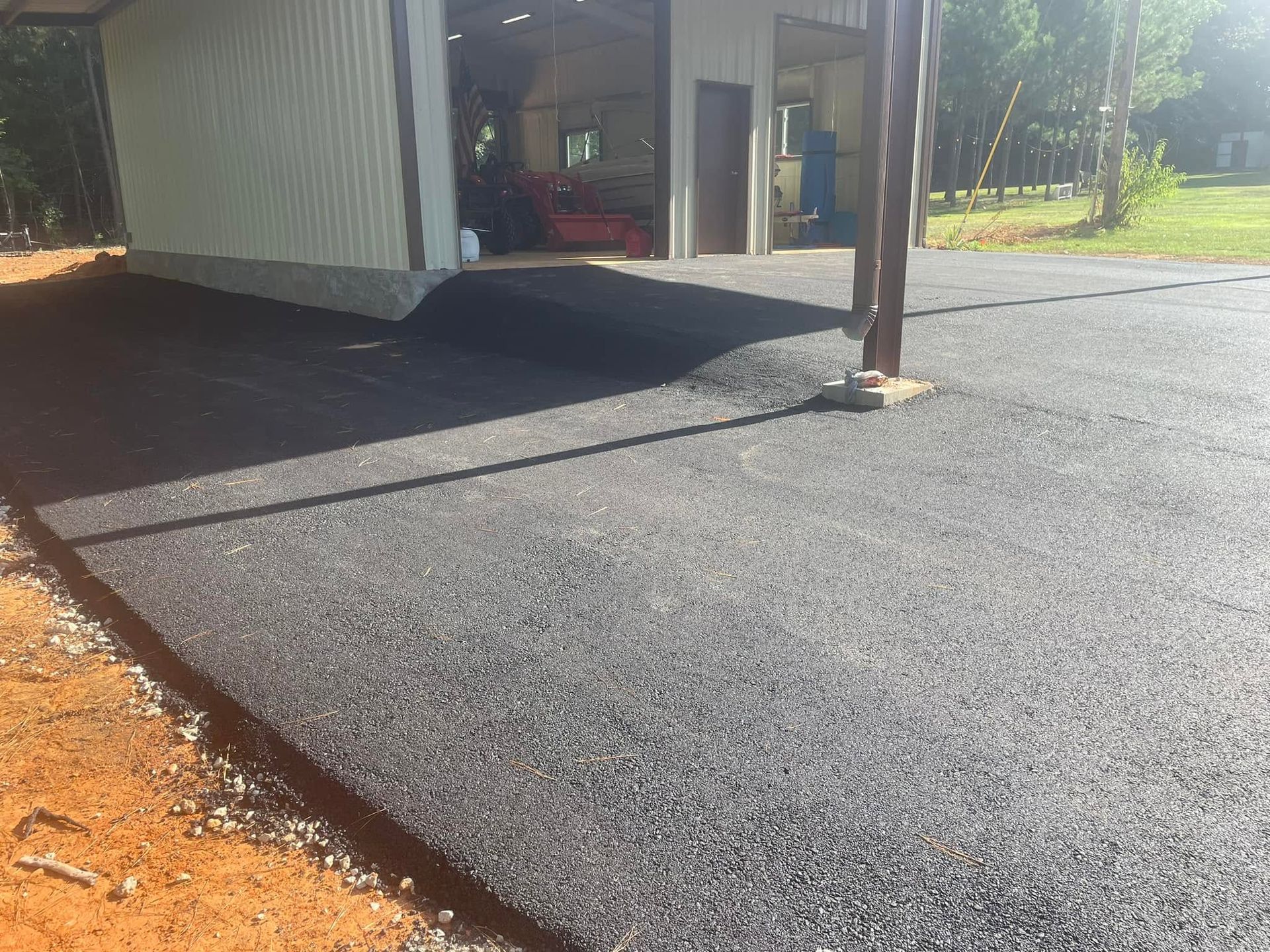 Freshly paved black asphalt around a metal-sided building and utility pole on an orange dirt lot.