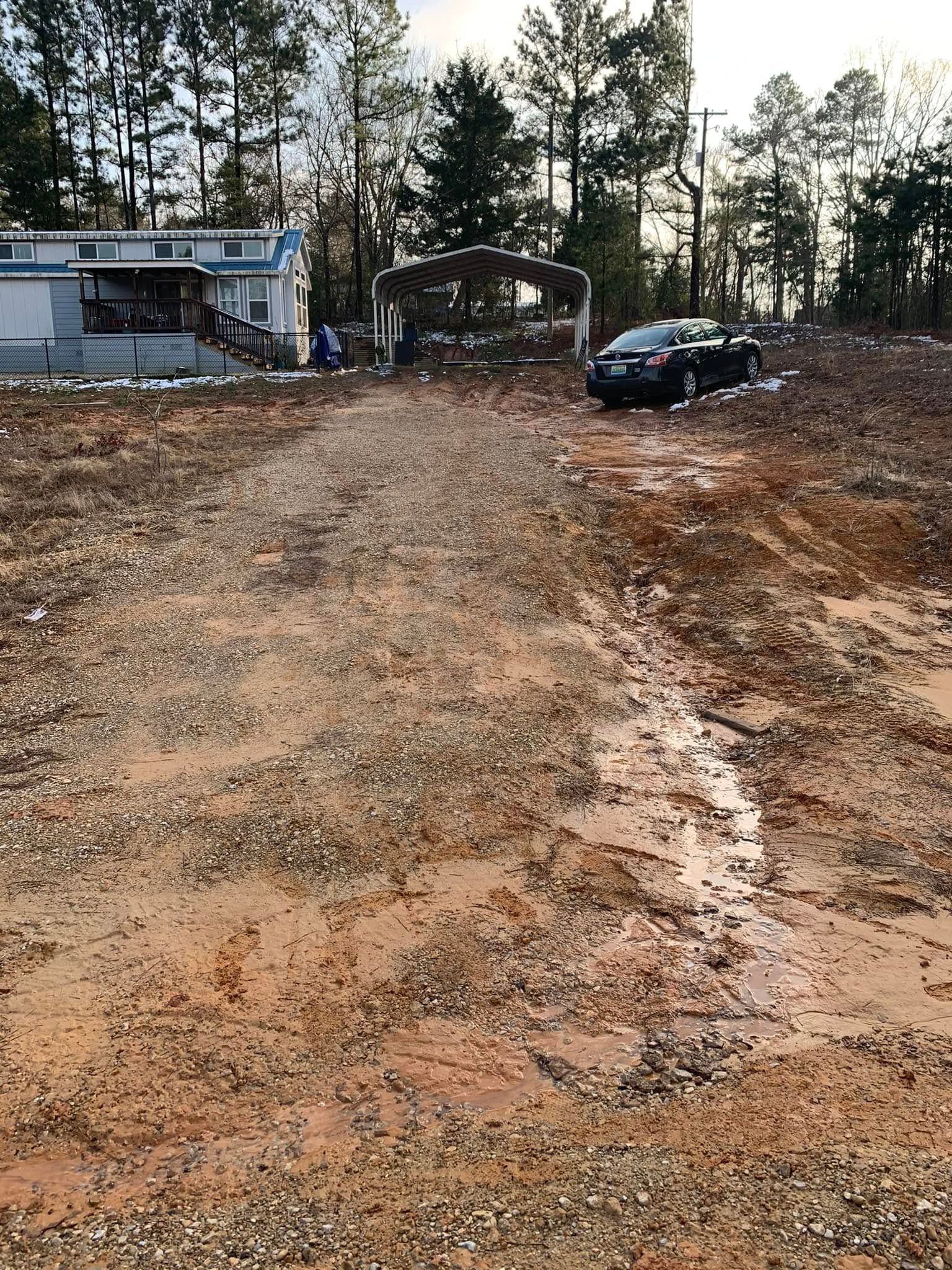 Wide dirt driveway leading to a wooded lot with a small building and a black car parked near the trees.