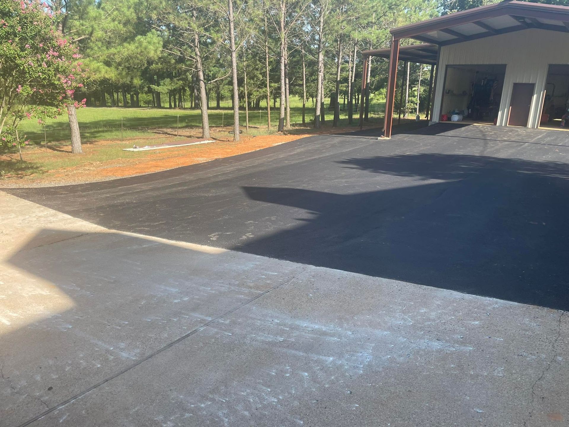 Concrete driveway beside a house with a carport, trees, and a grassy yard in bright sunlight
