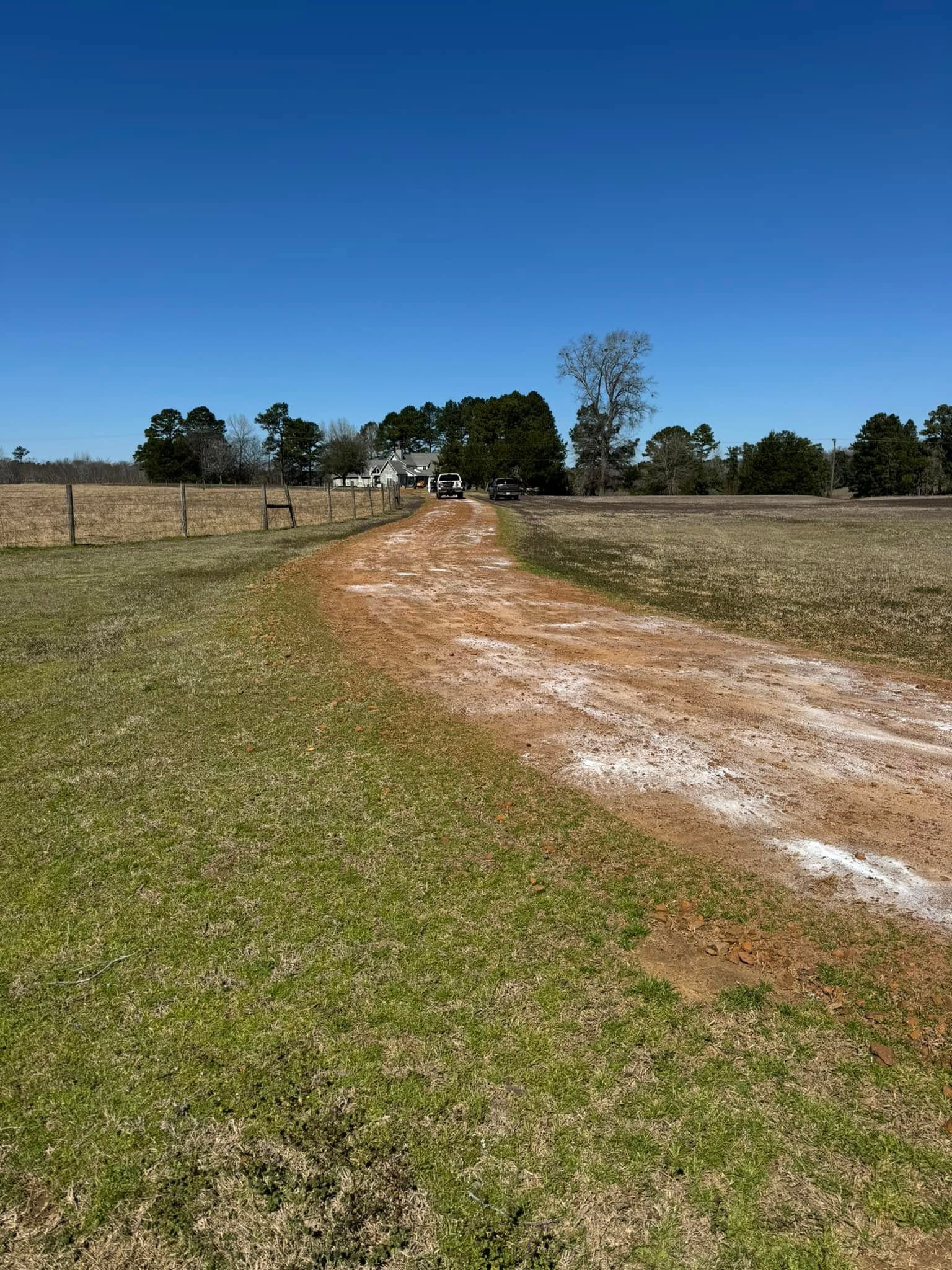 Dirt path through grassy field under a clear blue sky, with trees and a fence in the distance
