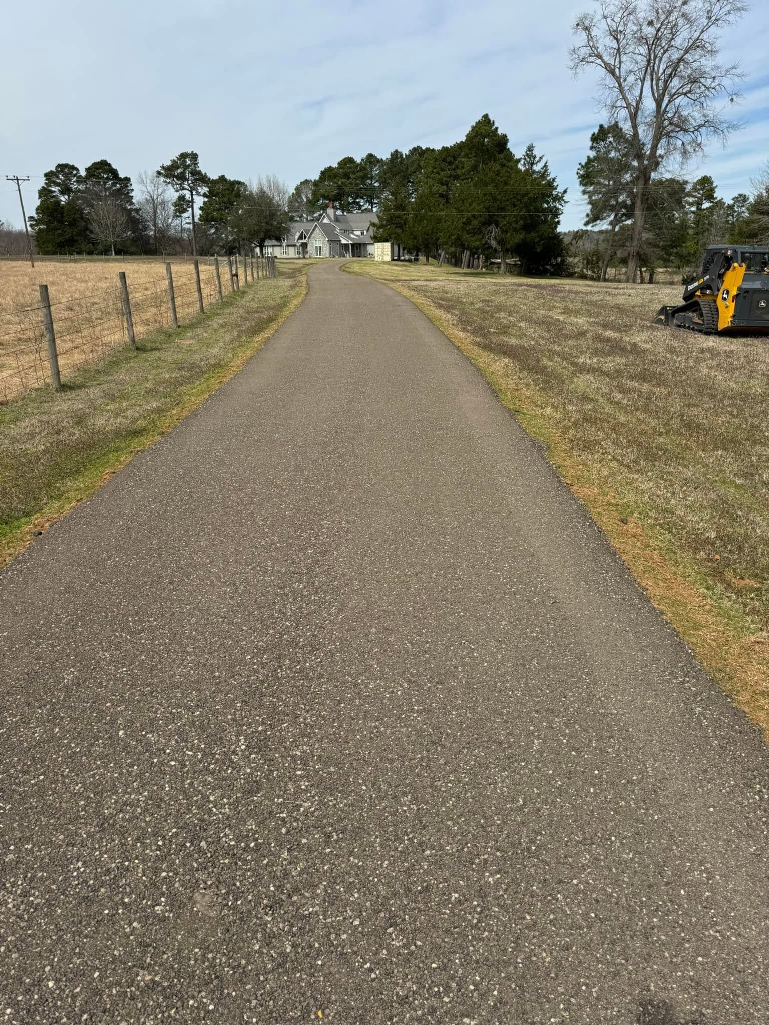 Gravel driveway leading to a house, with a fence on the left and trees under an overcast sky.