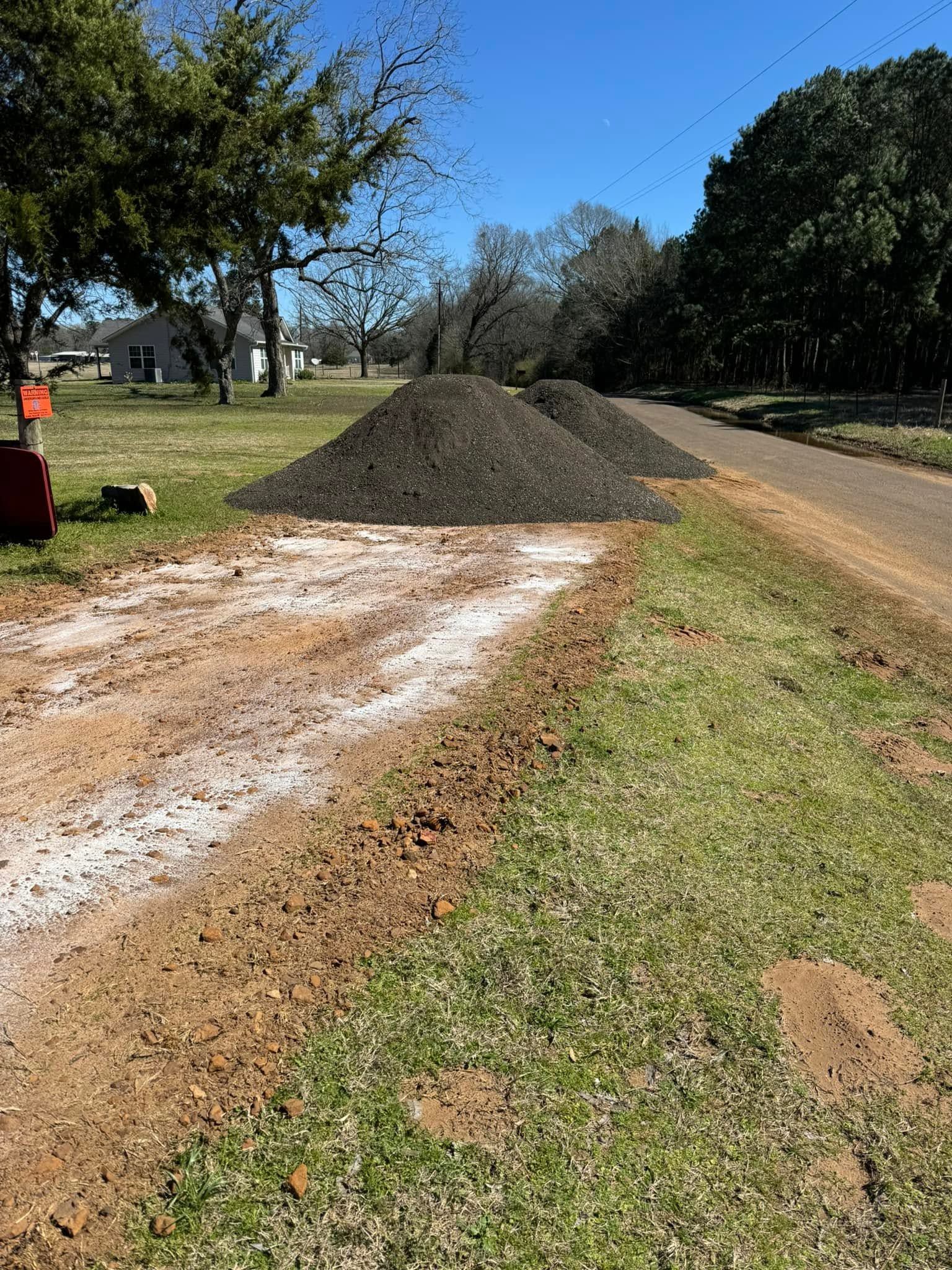 Gravel pile beside a dirt road with white powder on the ground, trees and grass along a sunny roadside