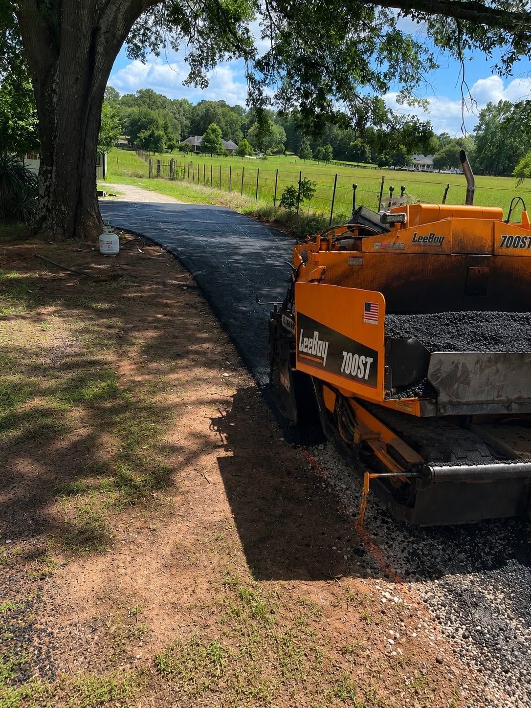 Orange construction machine on gravel path beside a tree-lined field under a blue sky