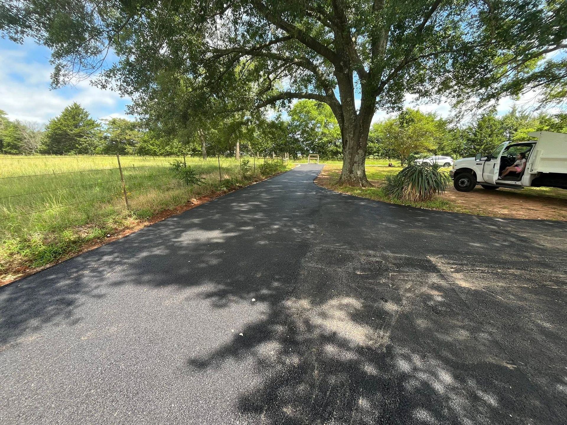 Tree-lined gravel driveway beside a grassy field, with a white truck parked on the right.