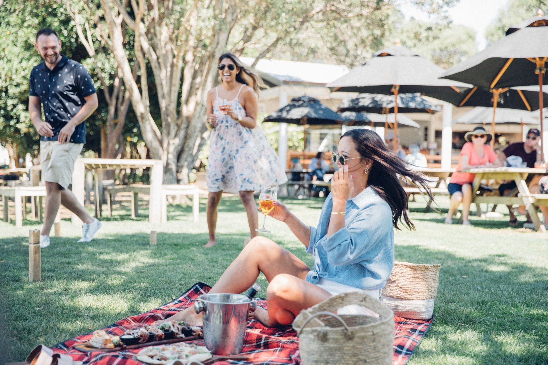 People enjoying a picnic outdoors. A woman sits on a blanket with food and drink; others stand nearby, and a man walks past smiling.
