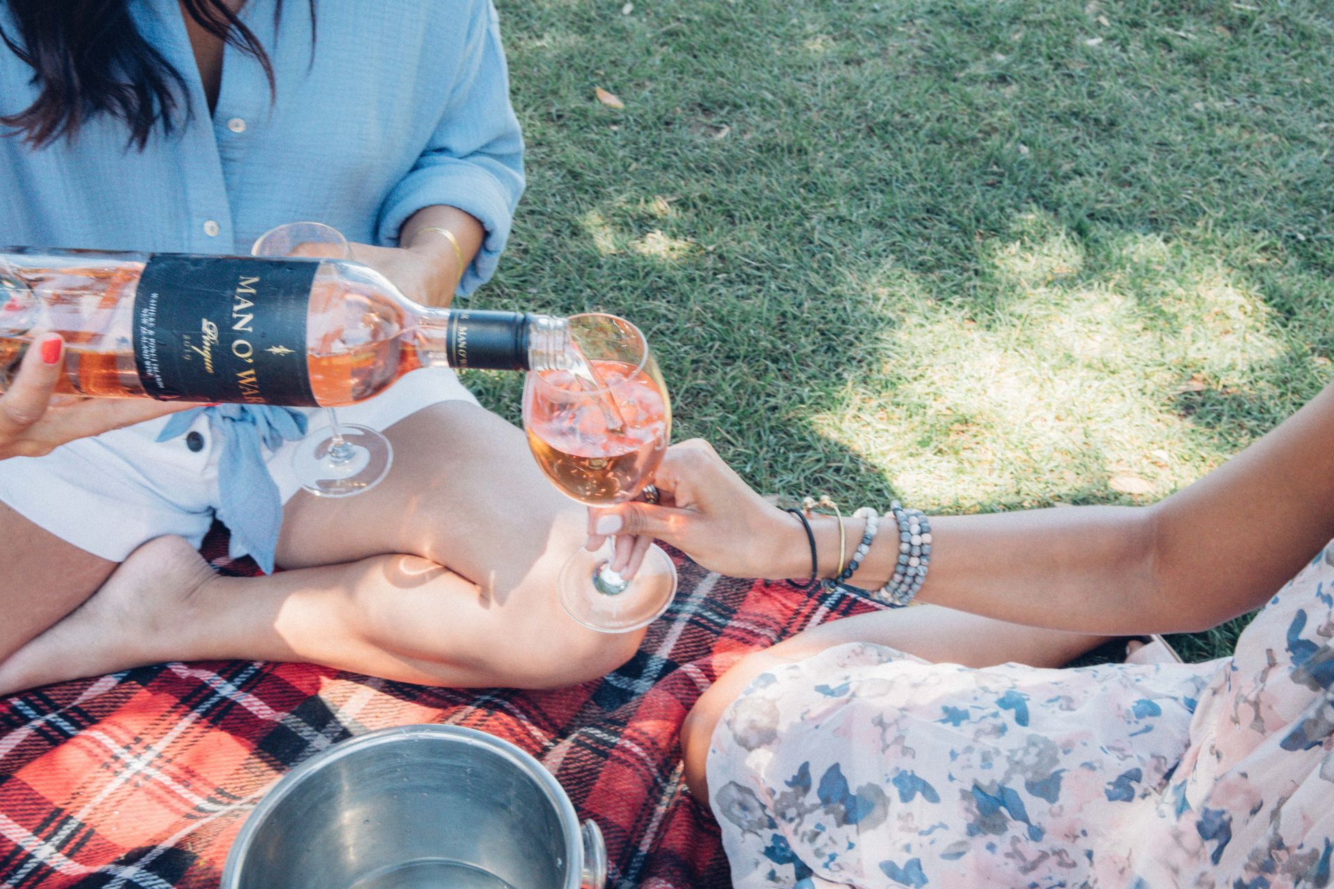 Person pouring Man O' War rosé wine into a glass during a picnic on a red plaid blanket in a grassy area.