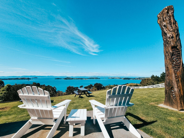 Two white Adirondack chairs on Waiheke Distiling Co lawn overlooking blue ocean to Corolmandel Peninsula under a clear sky