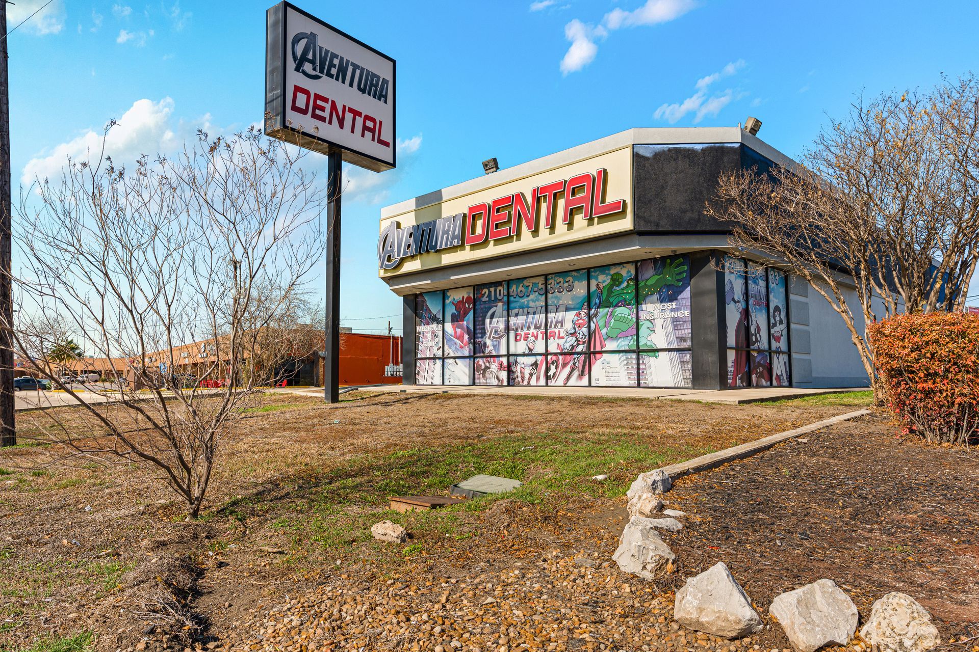 Dental office building with red signage and a freestanding sign, viewed from a landscaped roadside area