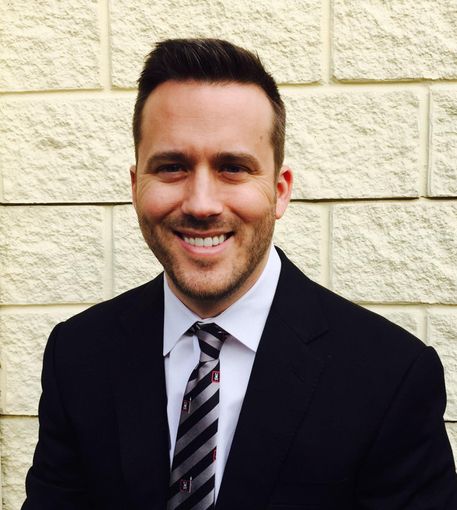 Smiling man in a black suit and striped tie standing in front of a stone wall