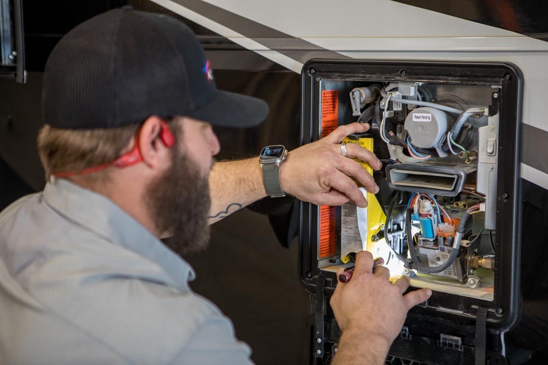 Person with beard working on an RV water heater in an open compartment. He is wearing earplugs and a cap.