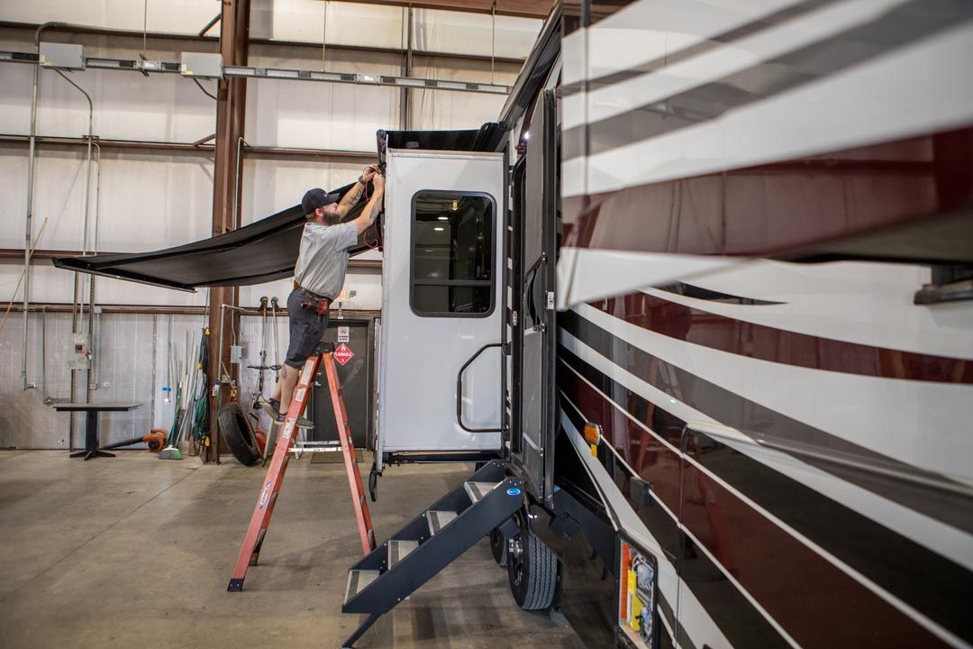 A technician on a ladder repairs the awning of a large recreational vehicle in a workshop.