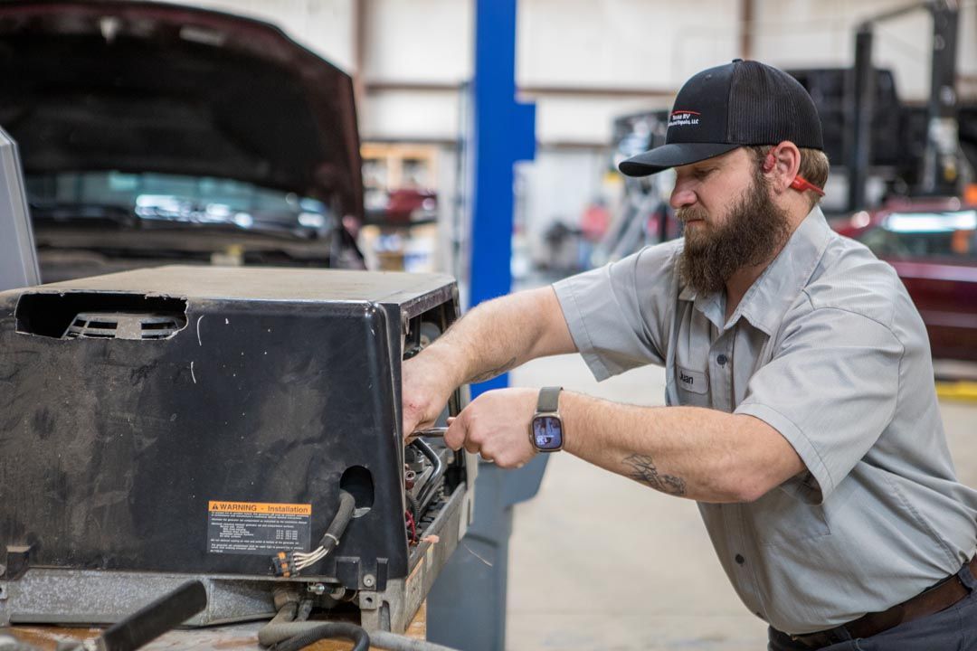 A mechanic in a grey uniform and baseball cap repairs a black metal machine component in an auto workshop.